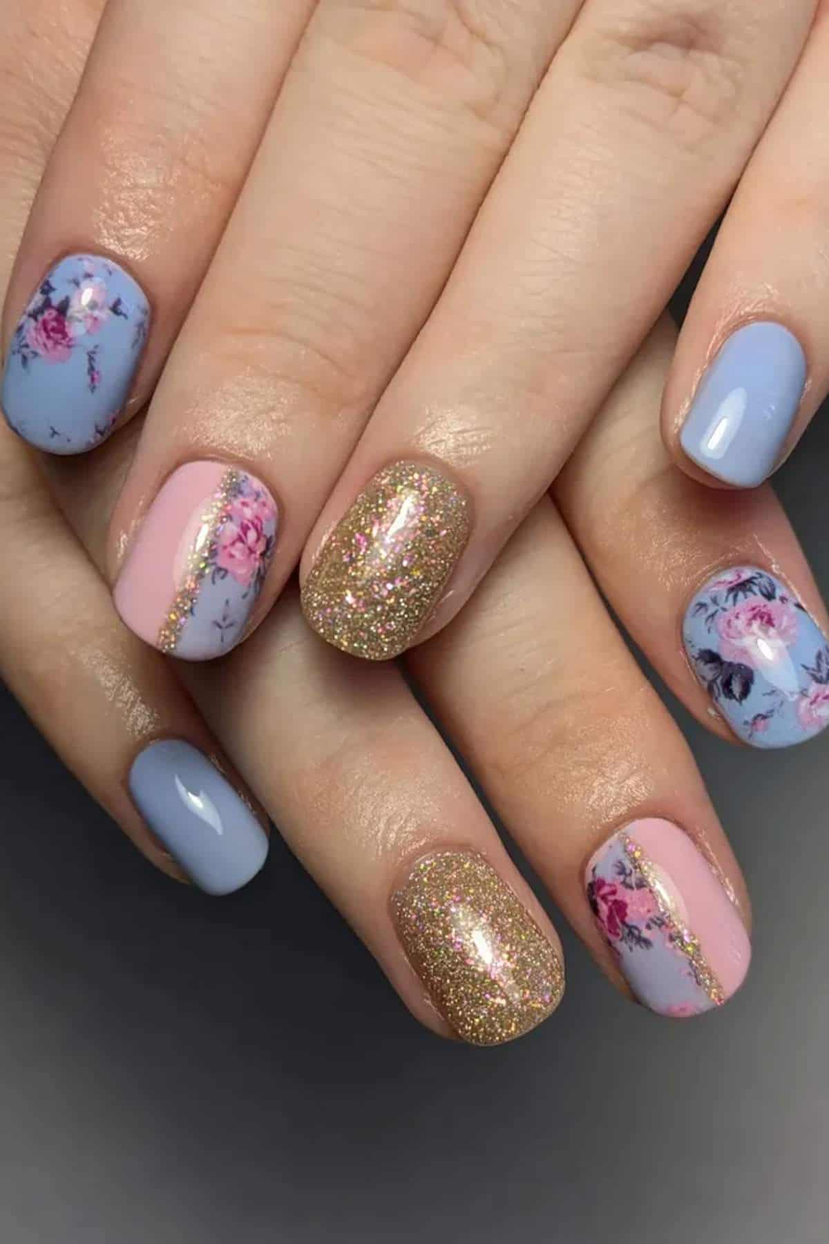 Hands with manicured nails featuring blue, pink, floral, and gold glitter designs, with some nails divided by a gold stripe.