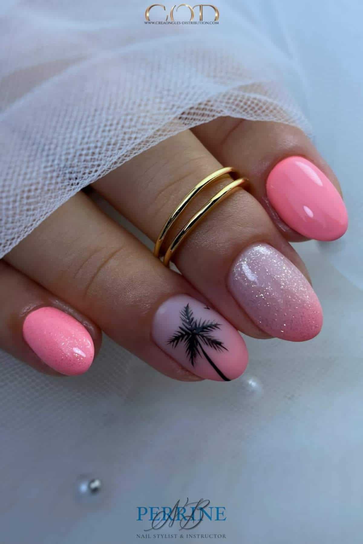 A hand with pink gradient manicure, one nail featuring a black palm tree design, resting on white fabric. The person is wearing two gold rings.