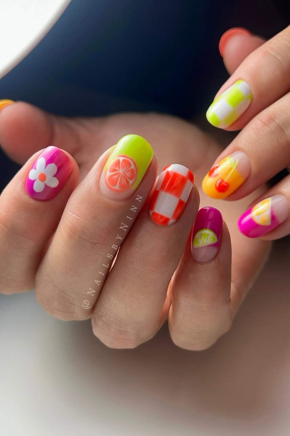 A hand with brightly colored nail art featuring designs of a flower, citrus slices, a cherry, and a red checkered pattern on different fingernails.
