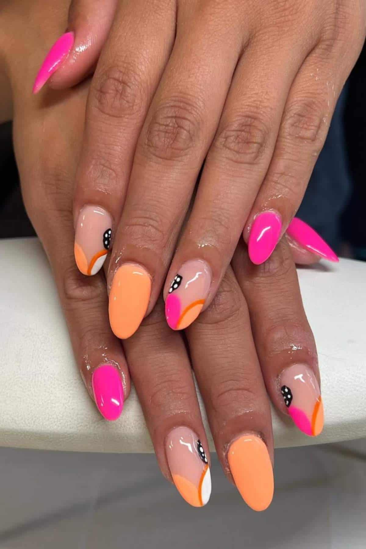 Hands with almond-shaped nails featuring a mix of bright pink, orange, and nude polish, some with abstract designs and small black and white dots.