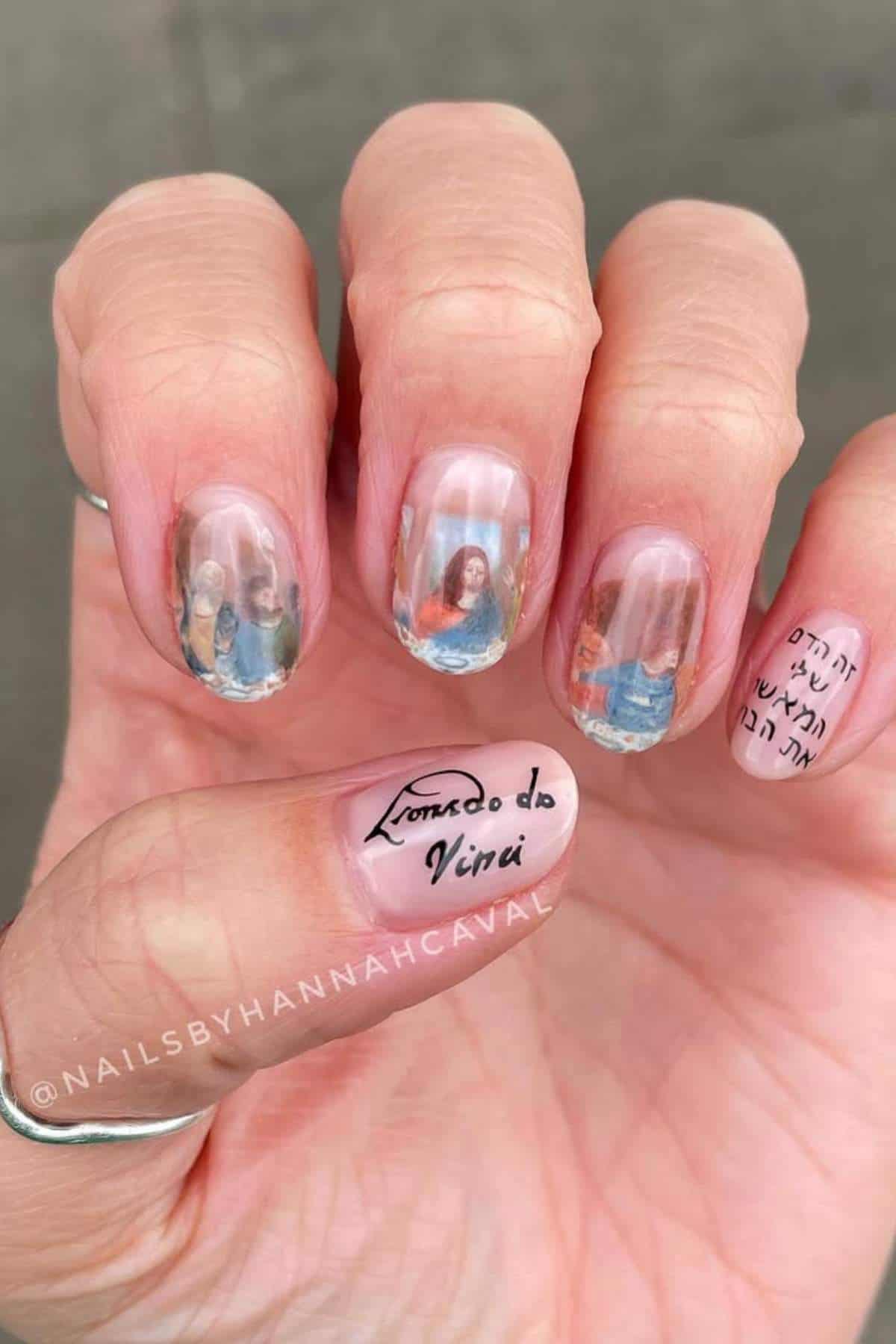 A hand with manicured nails featuring detailed photo art on four fingernails and handwritten text on the thumb and pinky nail.