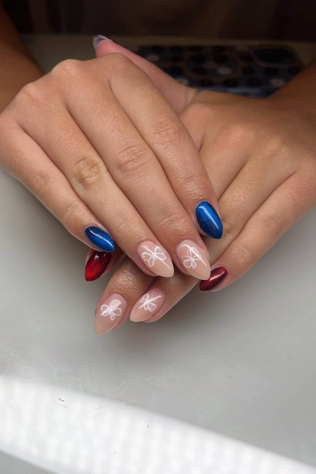 A pair of hands with manicured nails featuring metallic blue and red polish, and nude nails with white butterfly designs.