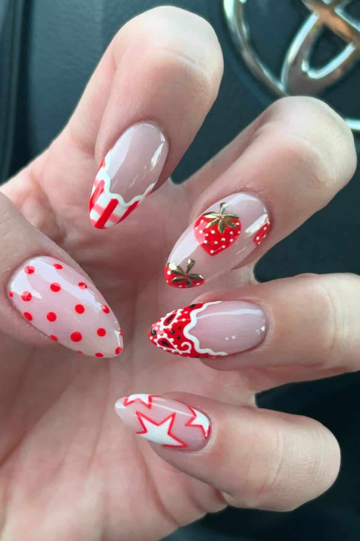 A hand with long almond-shaped nails featuring red and white designs, including strawberries, polka dots, stars, and wavy patterns, held in front of a car steering wheel.
