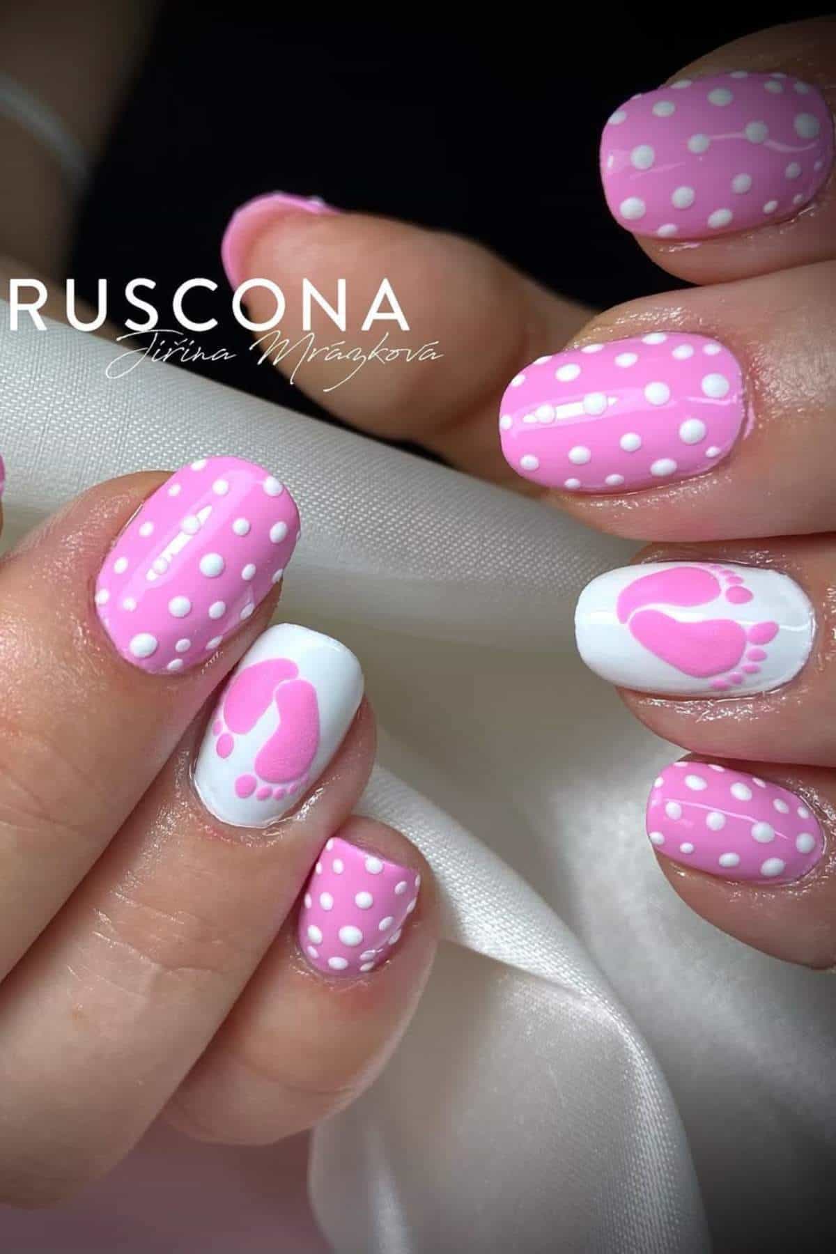 Close-up of hands with pink and white polka dot nail art; one nail on each hand features pink footprints on a white background.