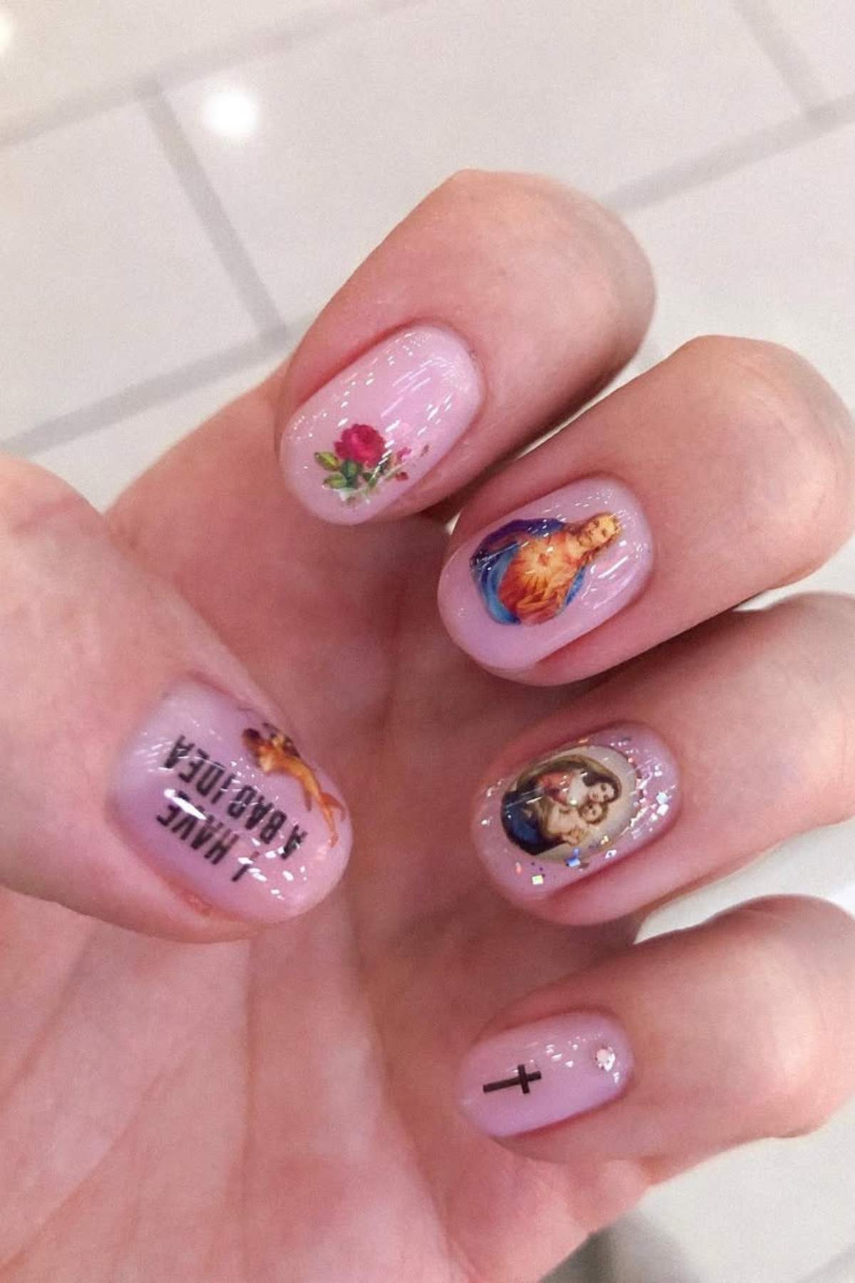 A hand with light pink manicure featuring small religious images, a rose, a cross, and text on the nails.