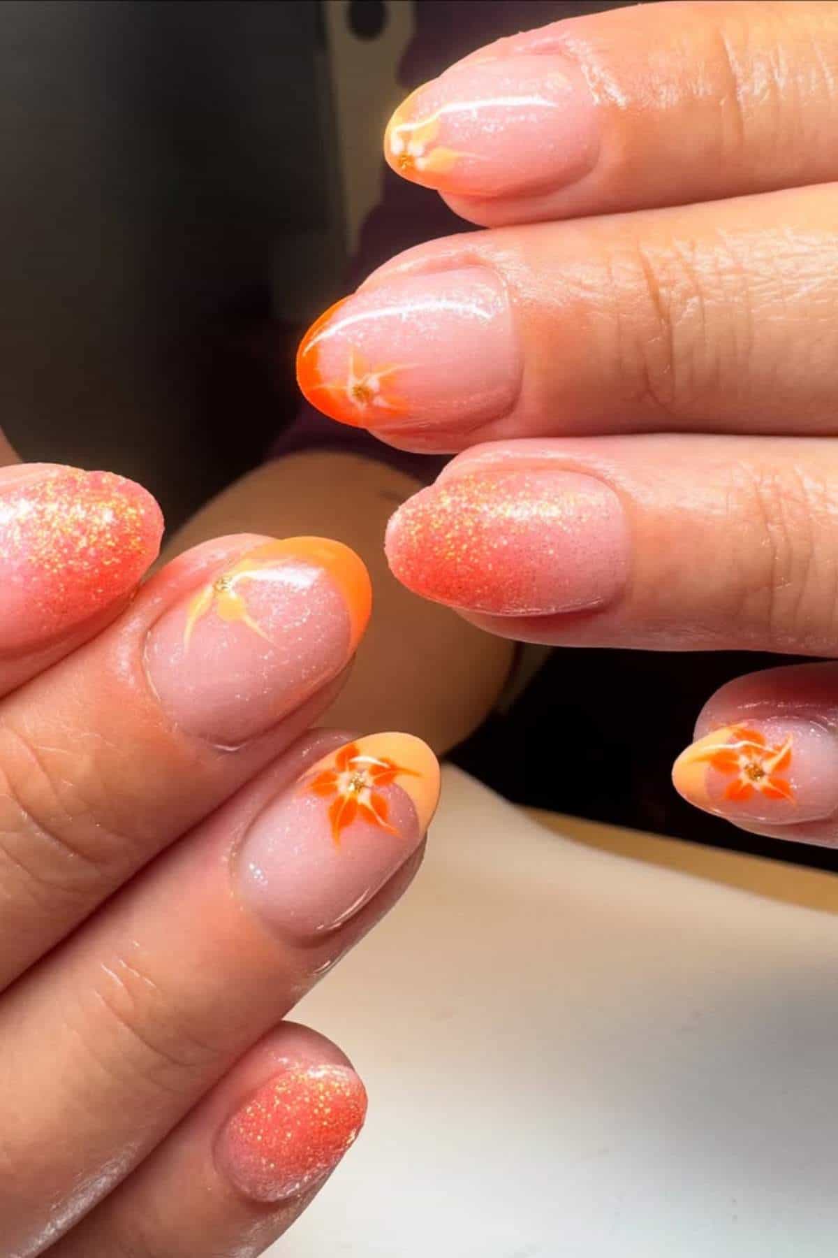 Hands with manicured nails featuring orange floral designs and glittery orange tips on a nude base.