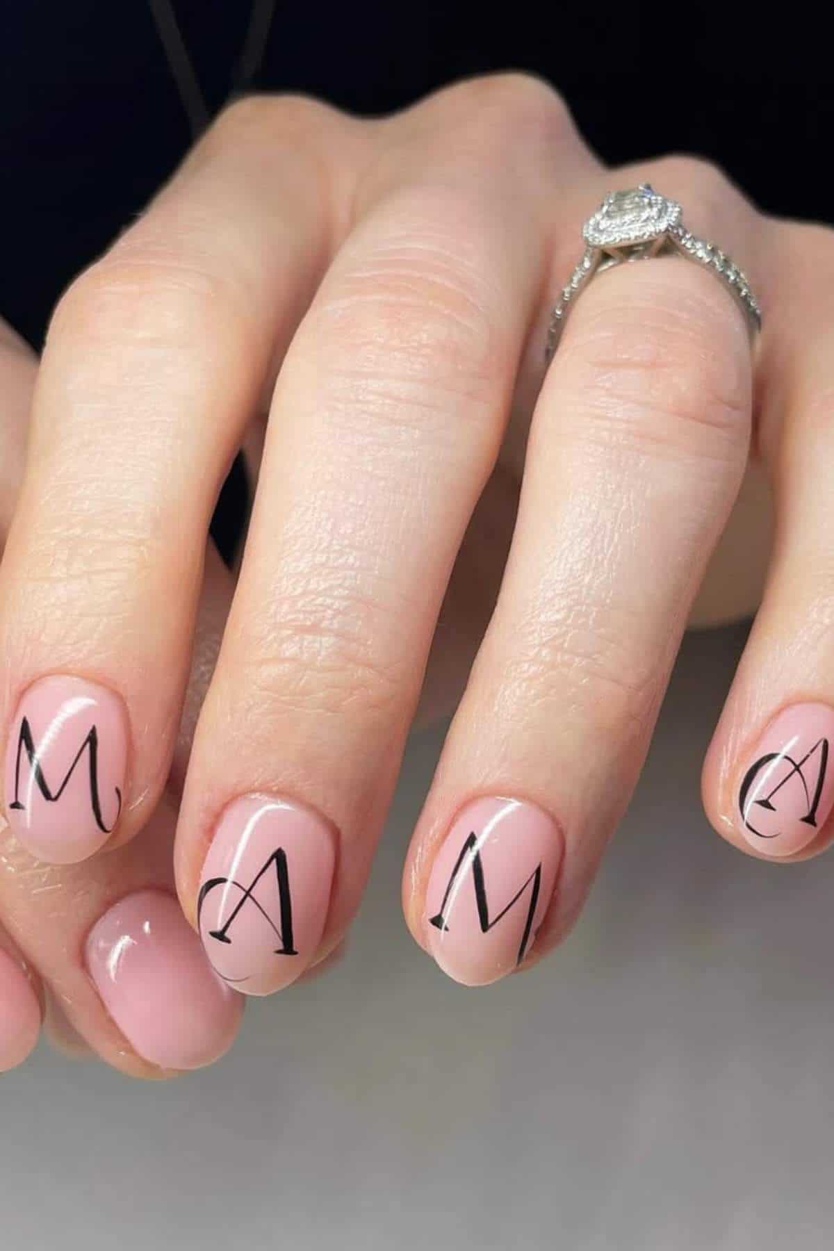A hand with light pink manicure; four fingernails have black letters spelling "MAMA." The person wears a diamond ring.