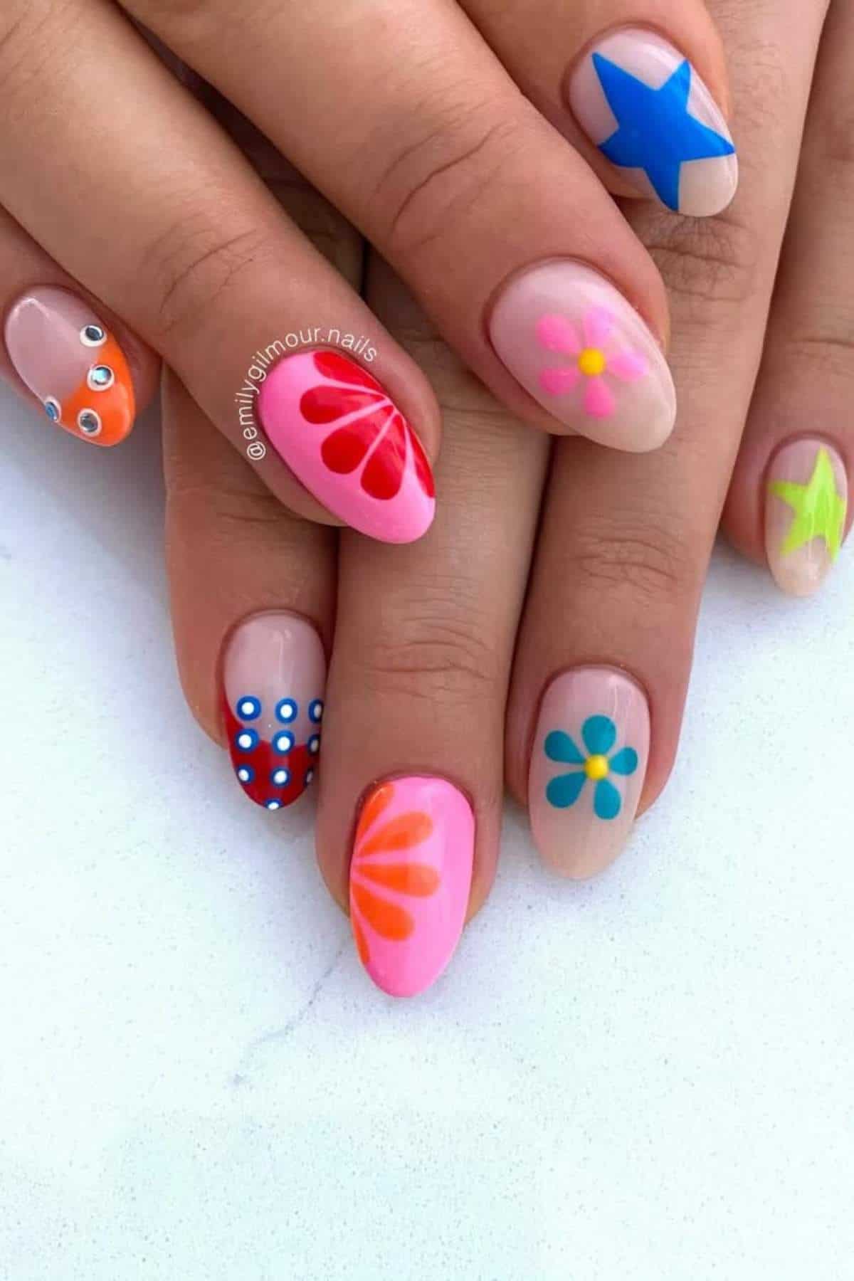 Close-up of hands with long nails featuring colorful designs, including flowers, stars, polka dots, and abstract patterns in bright shades of blue, pink, orange, and green.
