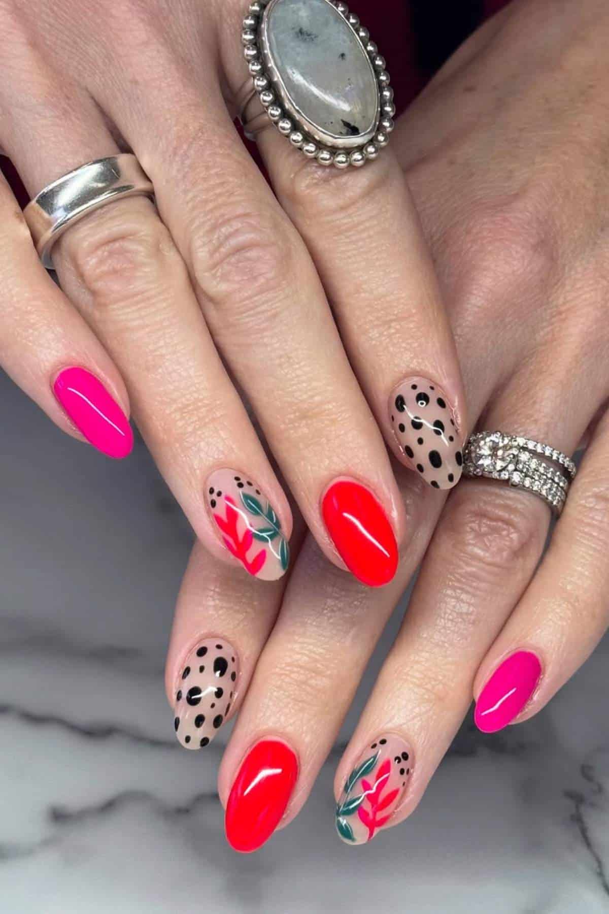 Hands with manicured nails featuring bright pink, red, black polka dots, and floral designs; person wears silver rings, including one with a large stone.