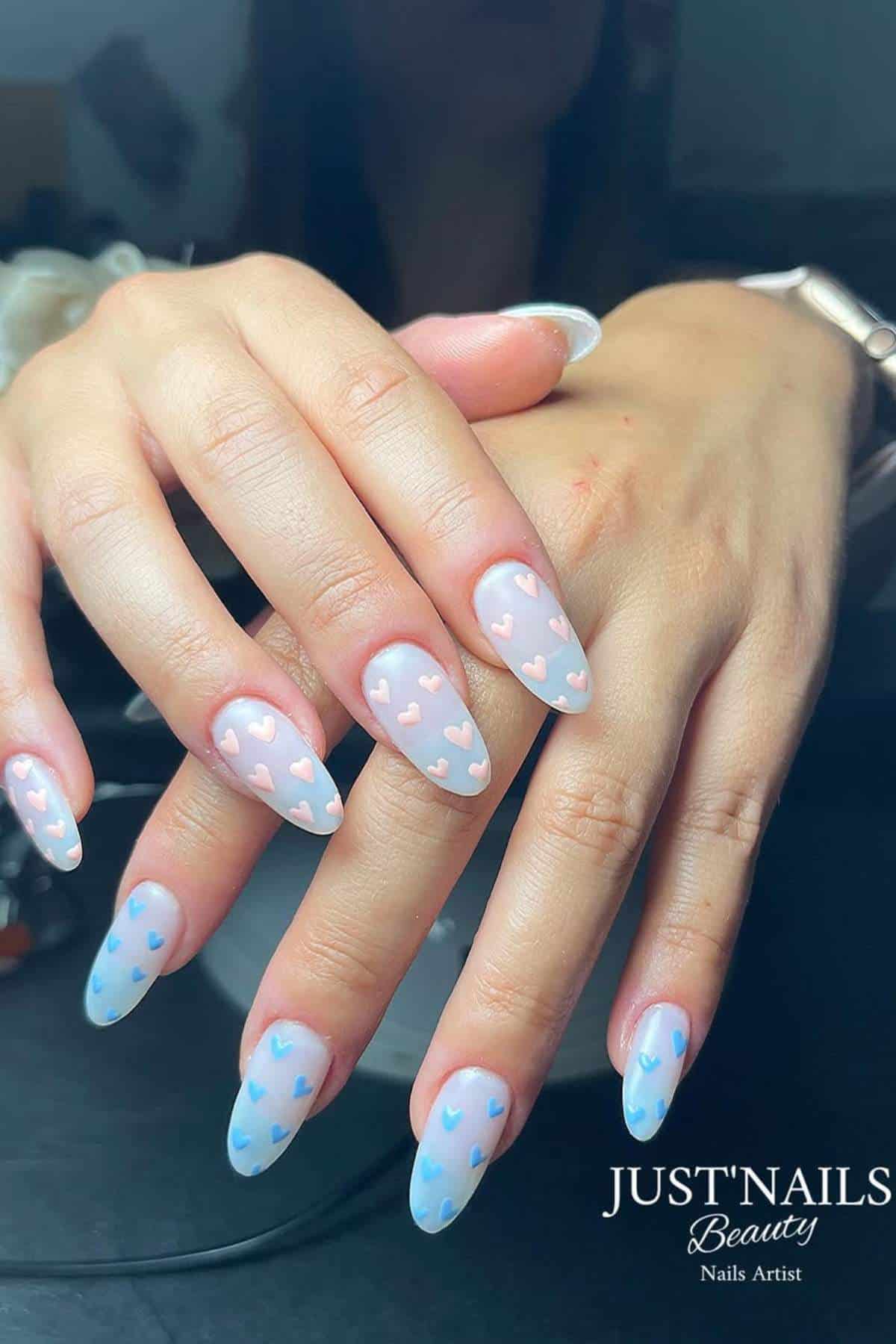Hands with long almond-shaped nails painted in a light pastel shade, featuring small pink and blue heart designs on each nail.