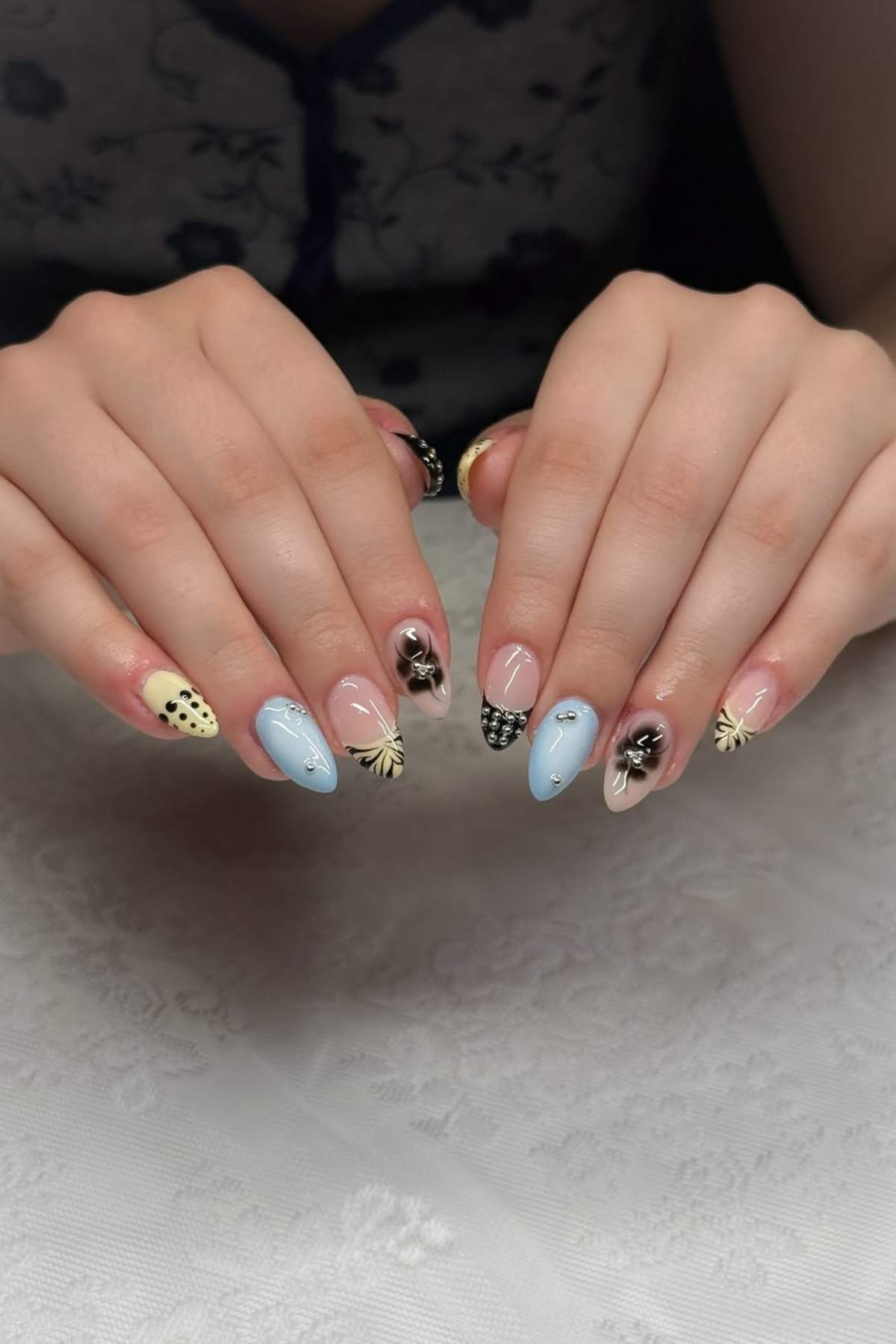 Two hands with manicured nails featuring a mix of pastel blue, black, and yellow designs, including polka dots, flowers, and rhinestone accents.