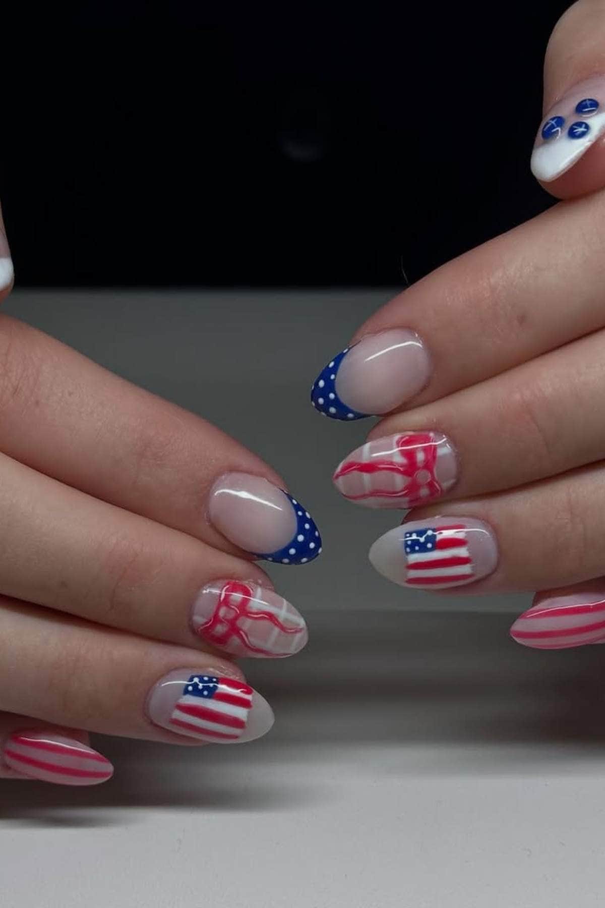 Hands with almond-shaped nails featuring American flag-themed nail art in red, white, and blue with stars, stripes, and polka dot designs.
