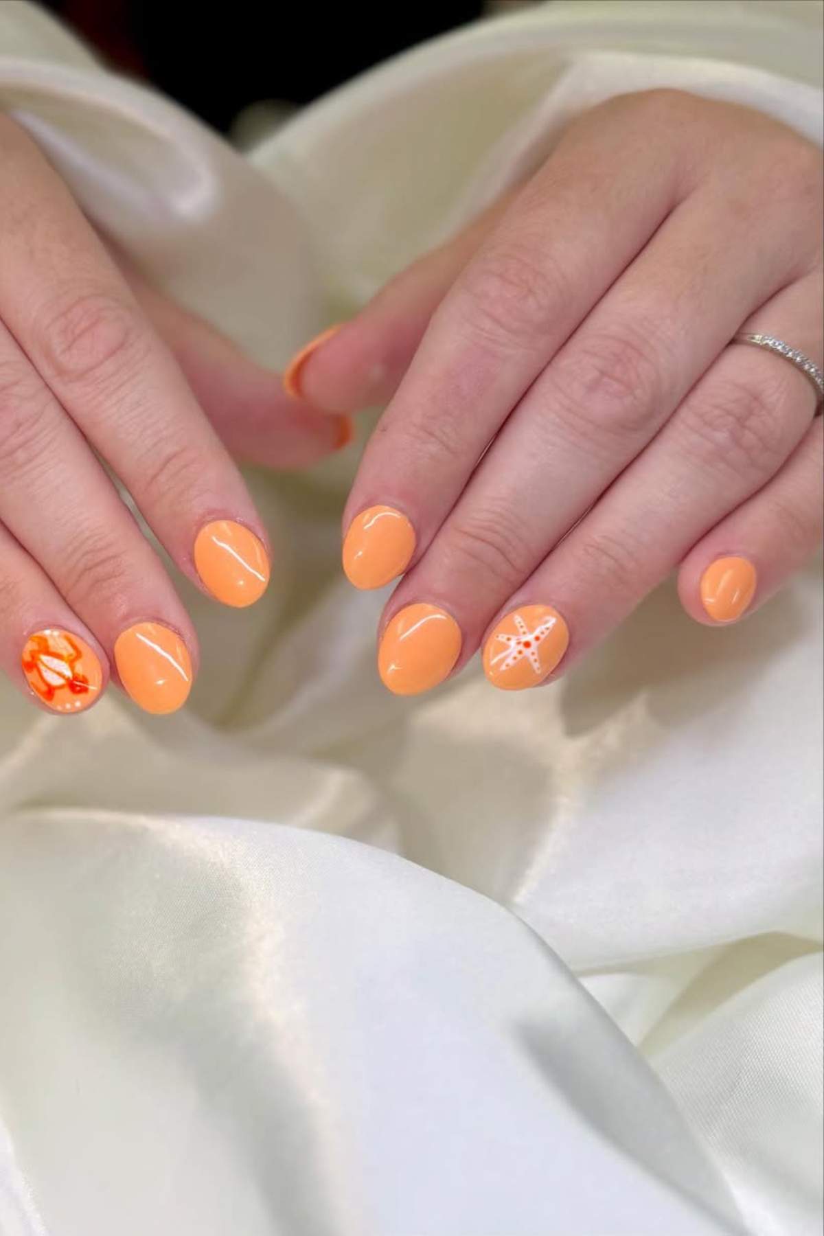 A pair of hands with almond-shaped nails painted orange; two feature white and orange art designs on a satin white fabric background.