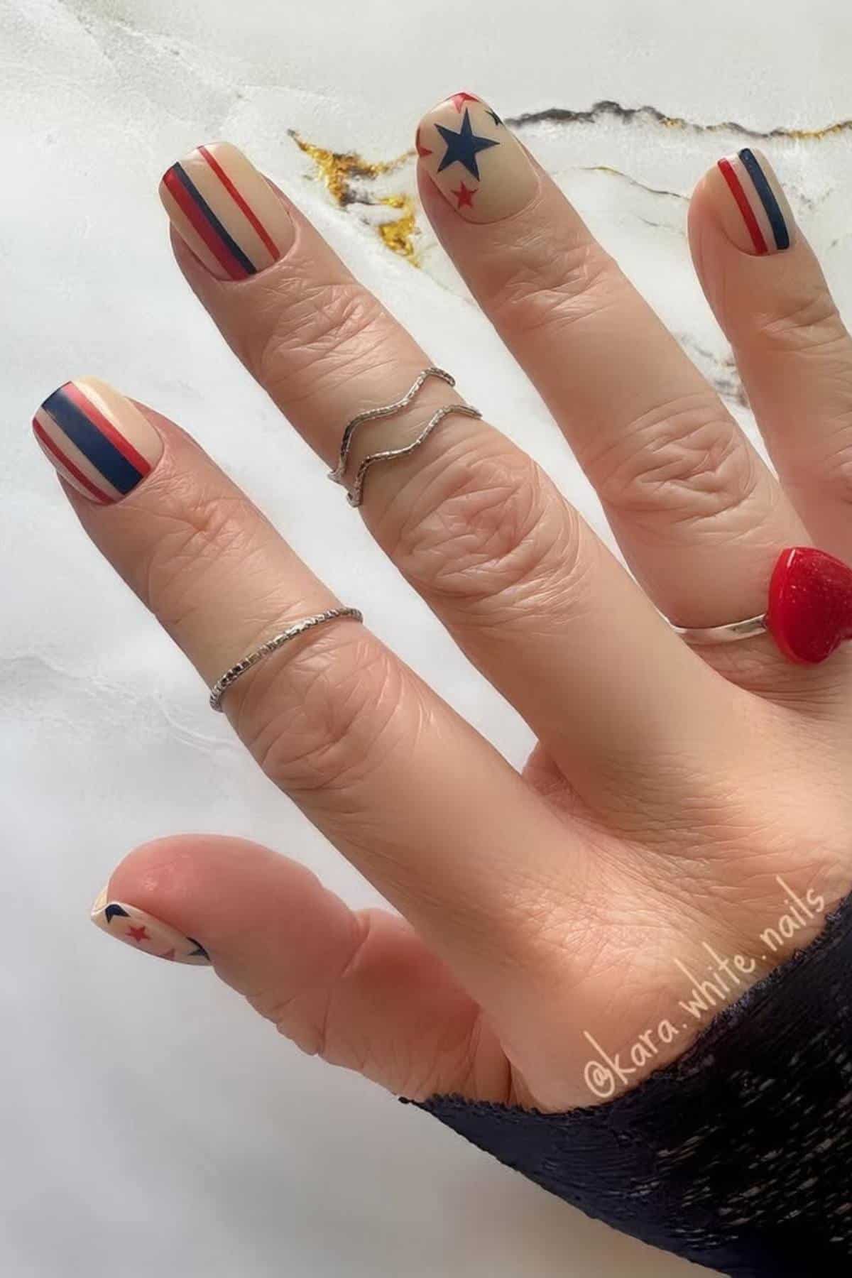 A hand with patriotic nail art featuring red, white, and blue stripes and stars. The person is wearing silver rings and a red heart-shaped ring.