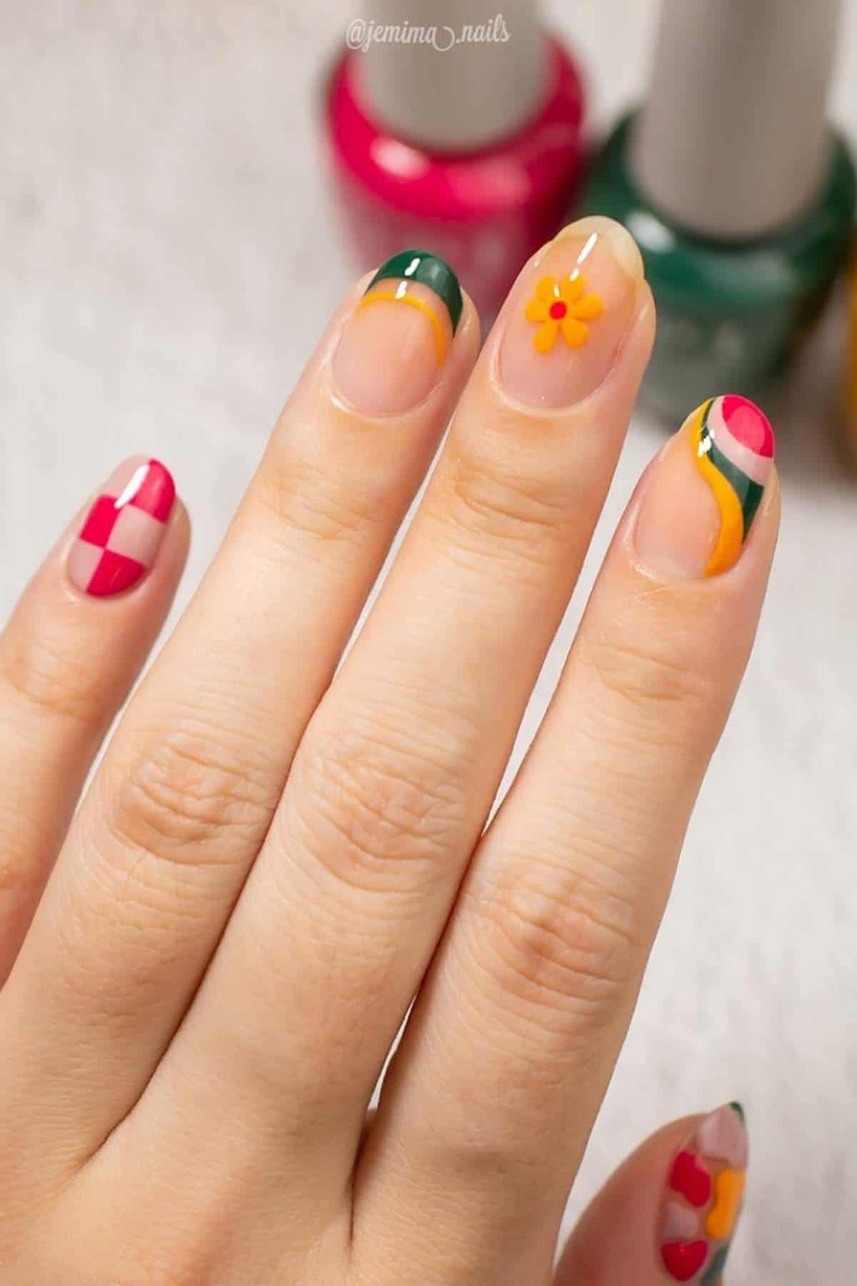 A hand with manicured nails featuring colorful, geometric and floral nail art designs in yellow, green, pink, and red tones. Three nail polish bottles are blurred in the background.