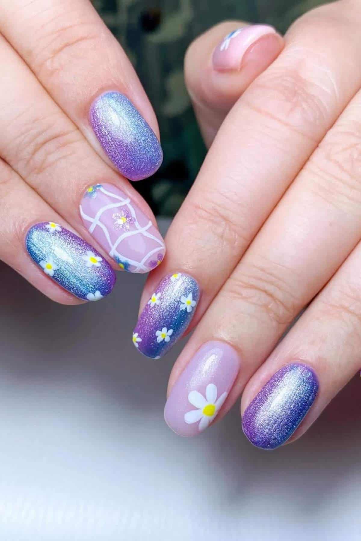 Close-up of hands with oval-shaped nails painted in purple and blue gradients, featuring daisy flower designs and white line patterns.