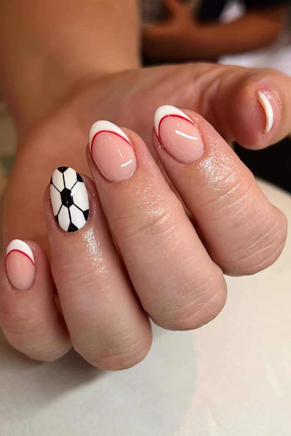 A hand with manicured nails; four nails have a French tip with a thin red line, and one nail is painted with a black and white soccer ball design.