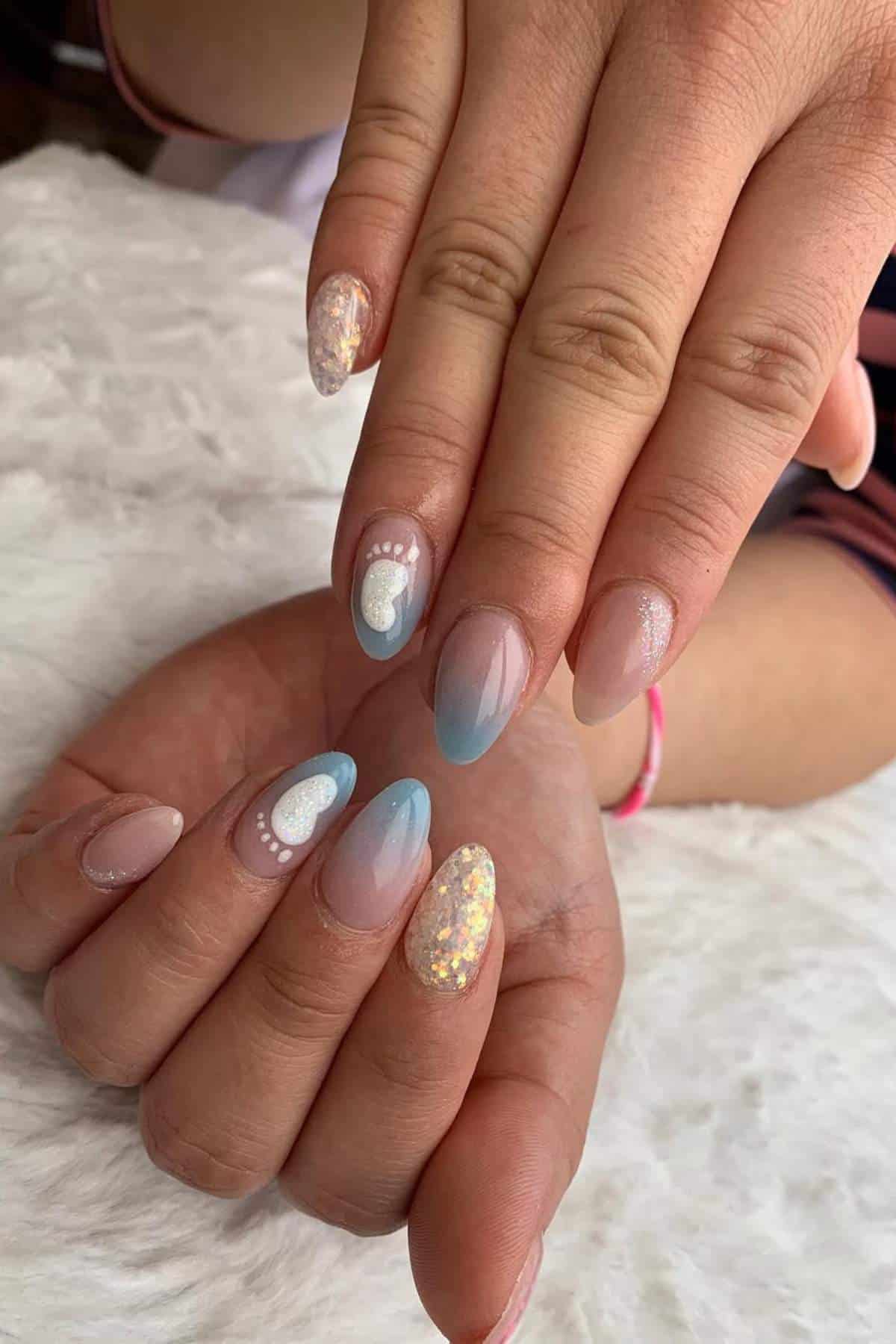 Hands with almond-shaped nails featuring pastel ombre, glitter, and white baby footprint designs, displayed on a white textured surface.