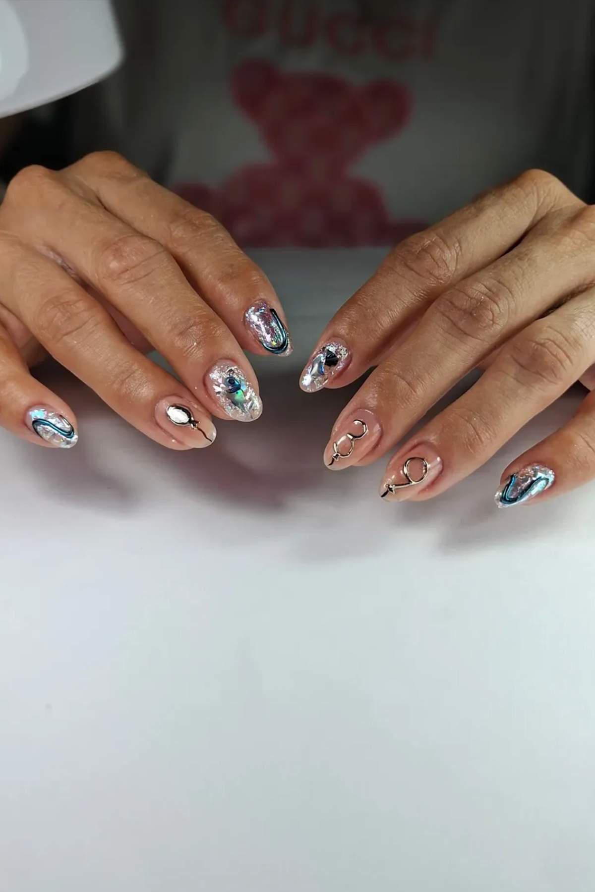 Hands with long, pointed nails featuring a shiny, metallic, abstract nail art design in blue, silver, and nude tones. The nails have a reflective, iridescent finish.
