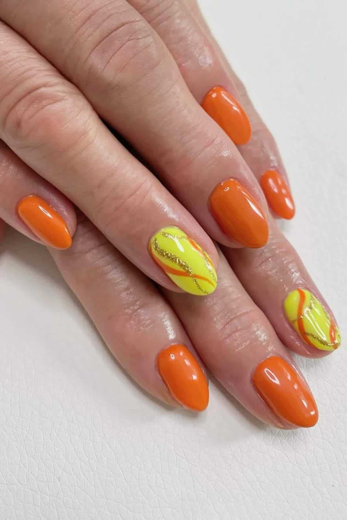 A hand with short, almond-shaped nails painted in glossy orange, with two accent nails featuring yellow, orange, and silver wavy designs.