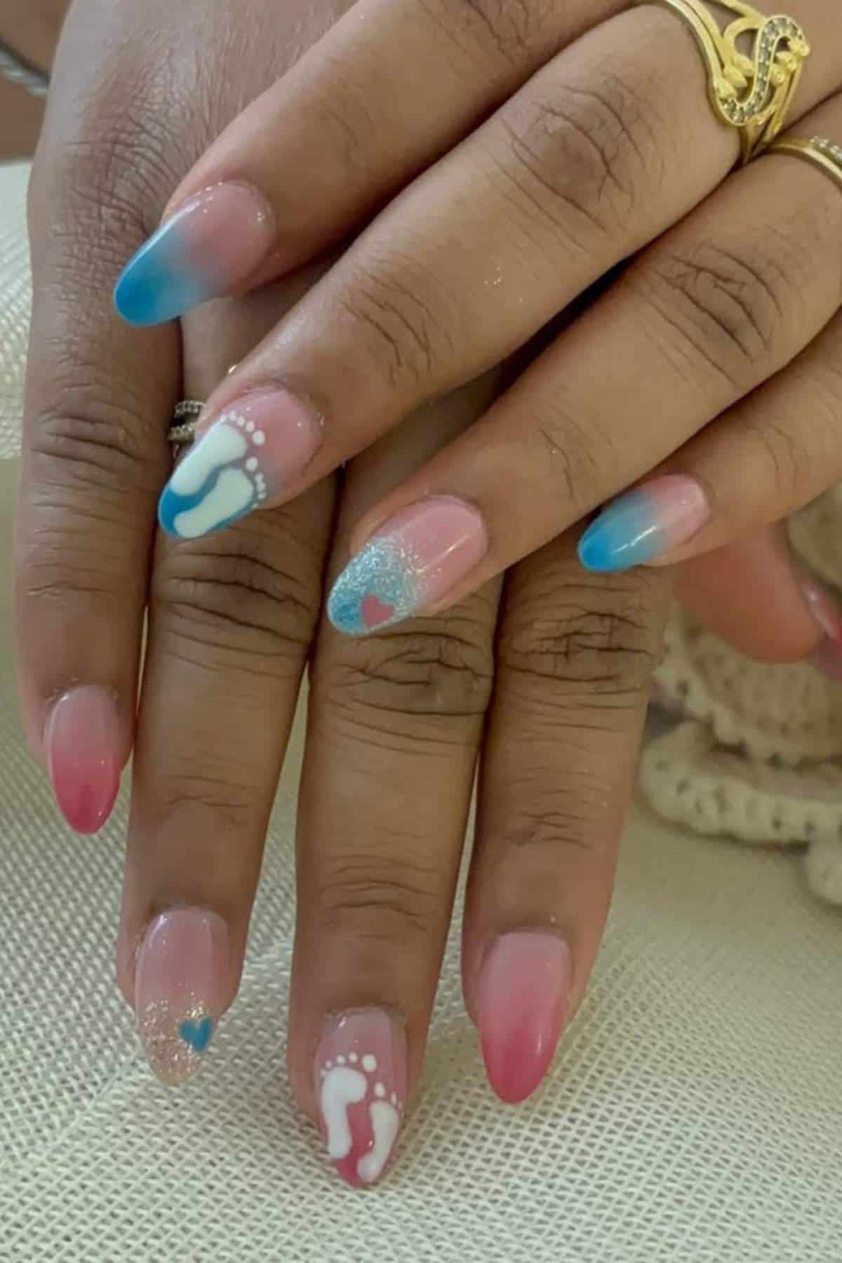 Hands with long, almond-shaped nails featuring pink and blue gradient polish, white baby footprints, glitter, and heart designs. Person is wearing gold rings.