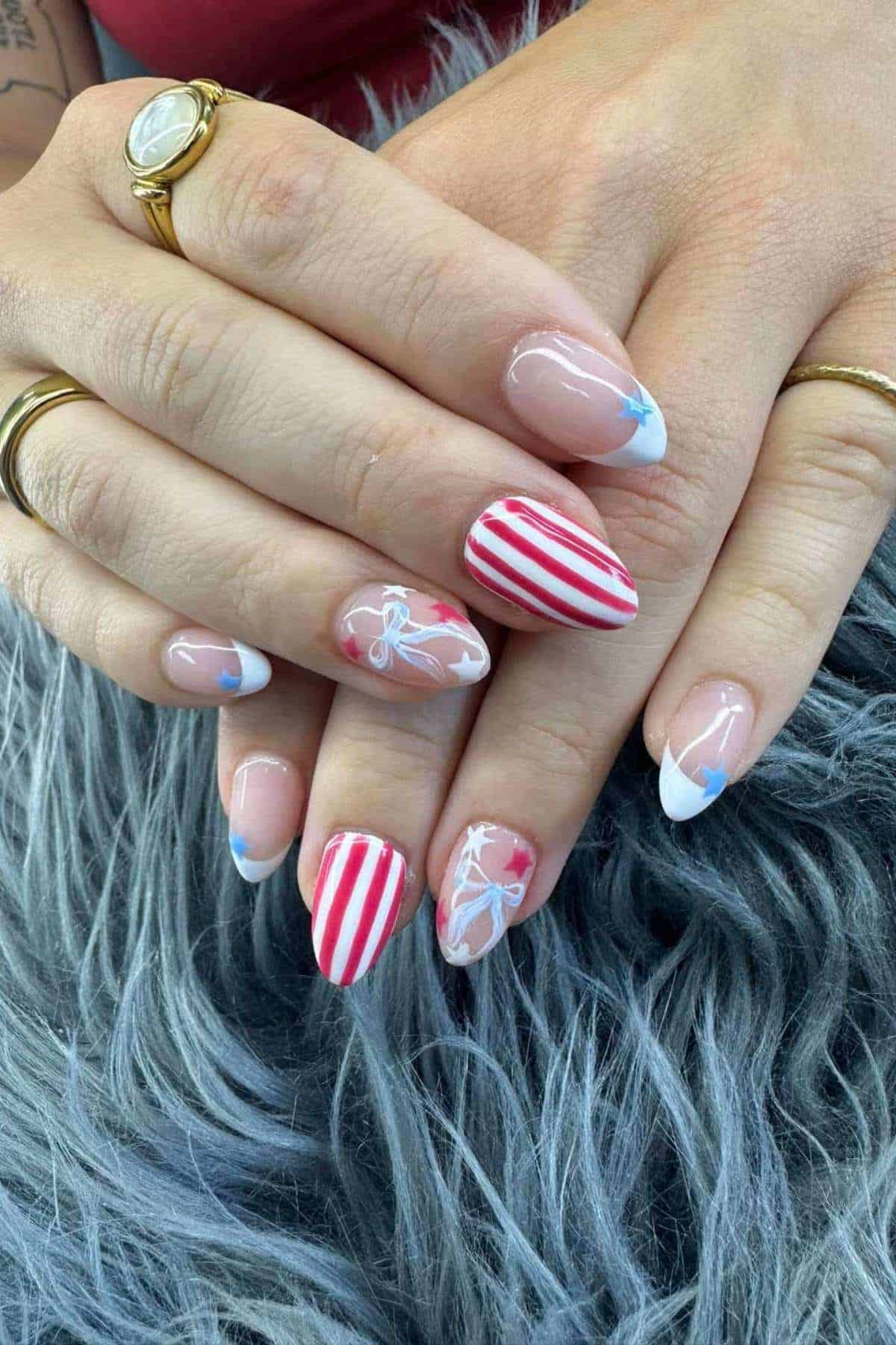 Hands with long almond-shaped nails featuring red and white stripes, blue tips, and white bow designs on a gray fur surface.