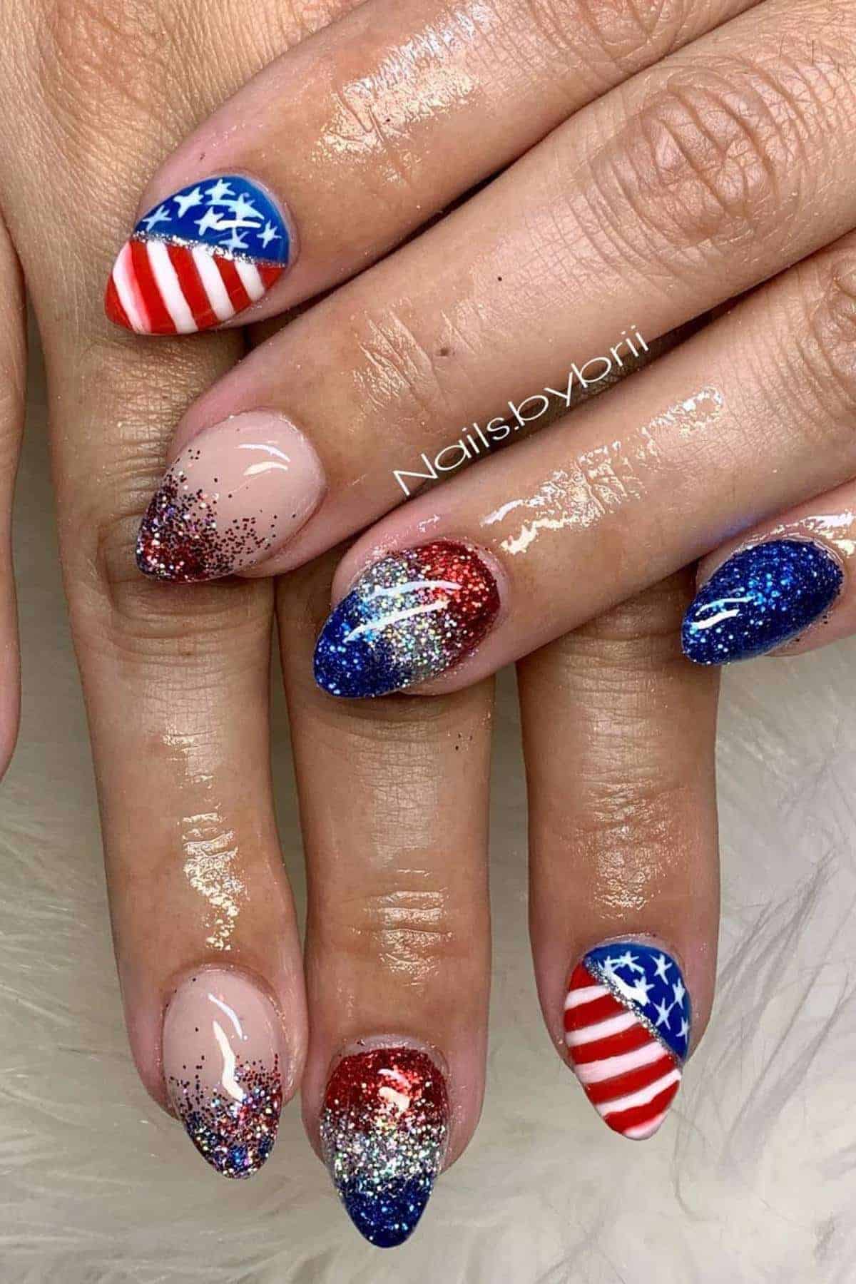 Hands with almond-shaped nails featuring American flag designs on some nails and red, blue, and silver glitter gradients on others.