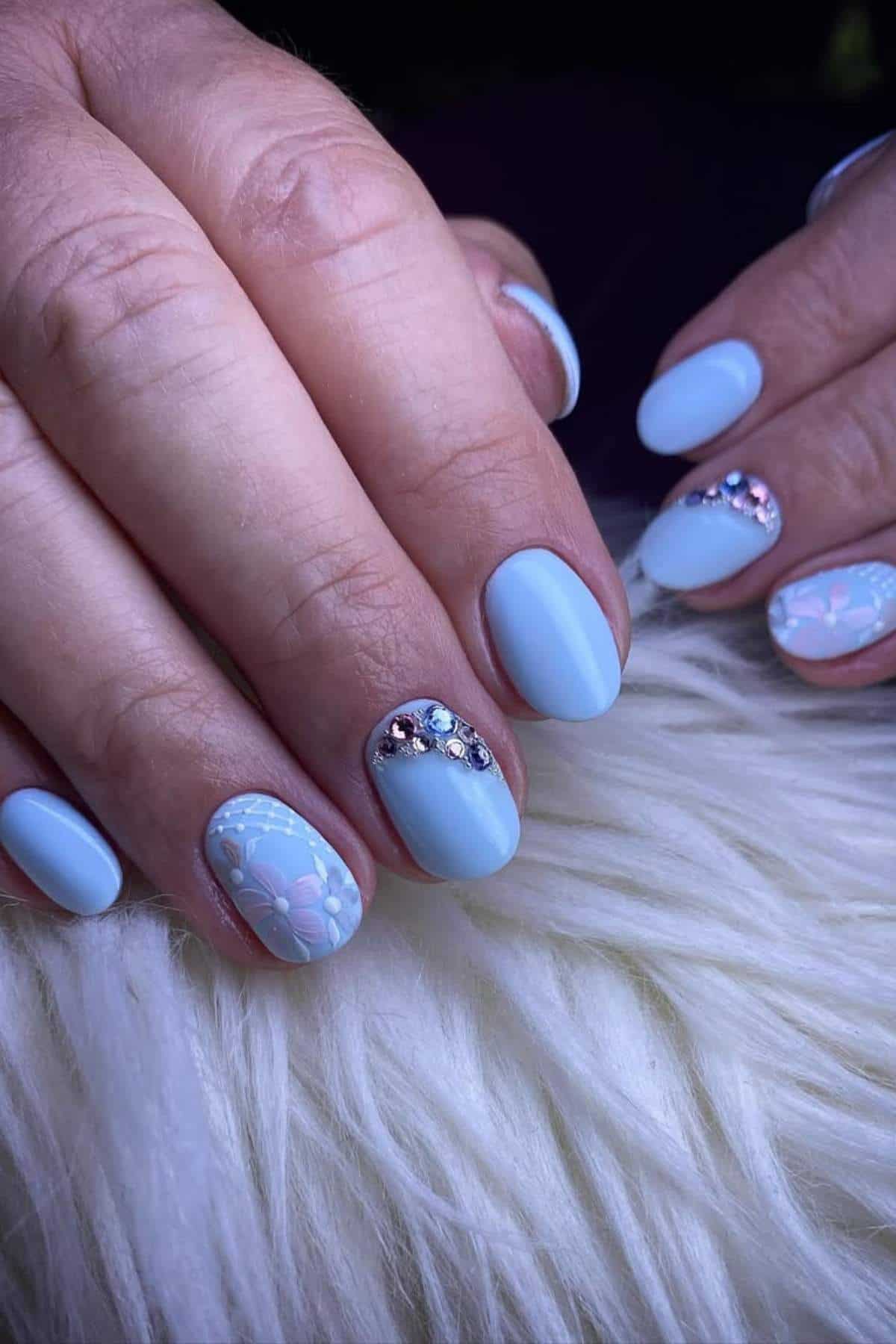Close-up of hands with light blue matte nail polish, floral accent nail art, and rhinestone embellishments on a white furry surface.