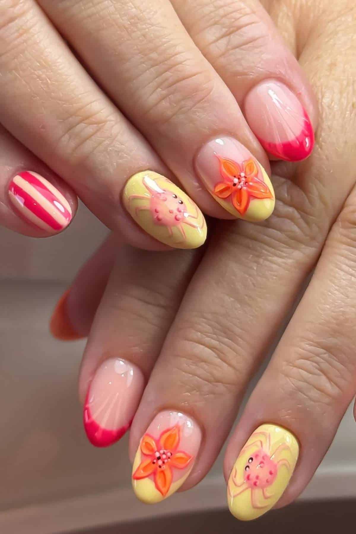 Hands with manicured nails featuring pink tips, yellow and pink floral designs, and one nail with pink stripes on a light background.
