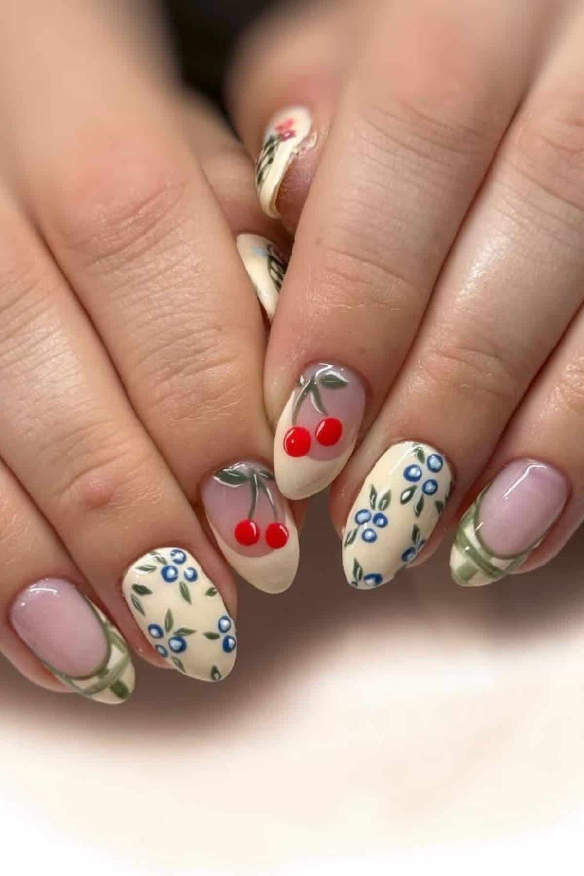 Hands with almond-shaped nails featuring nail art of blue flowers, red cherries, and green lines on a cream and clear background.