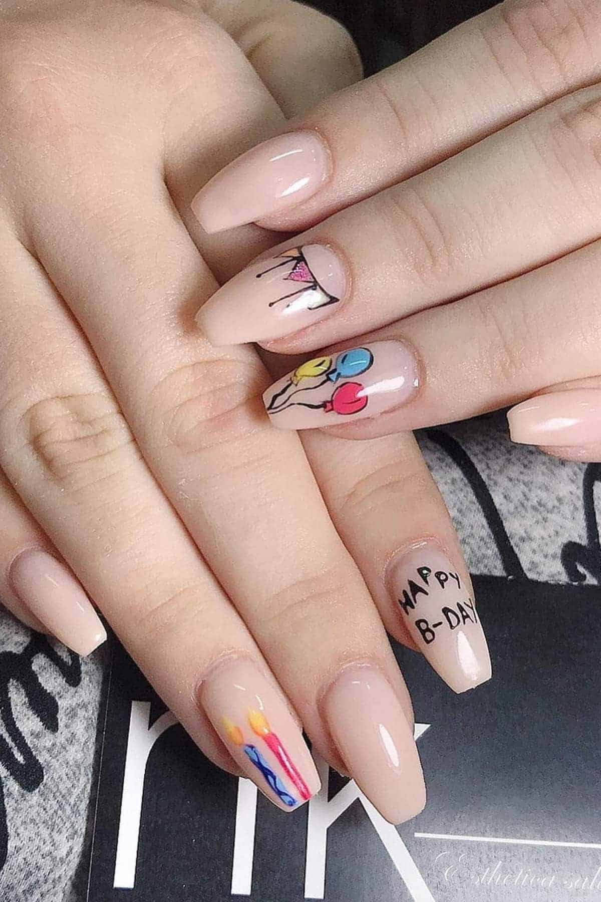 A hand with nude-colored nails, featuring birthday-themed nail art including balloons, candles, "HAPPY B-DAY" text, and a slice of cake.