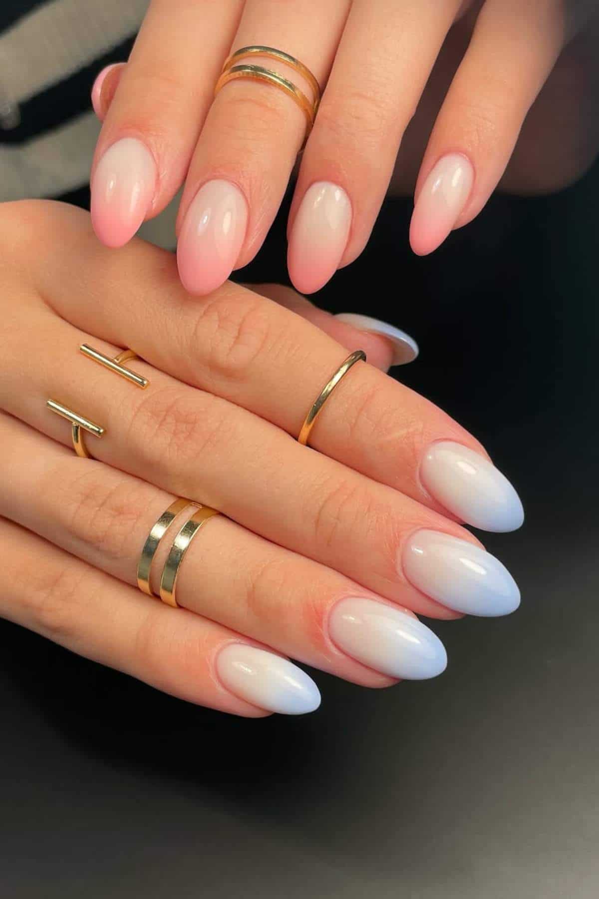 Close-up of hands with almond-shaped nails in a pink to white ombre design, wearing several gold rings on the fingers.