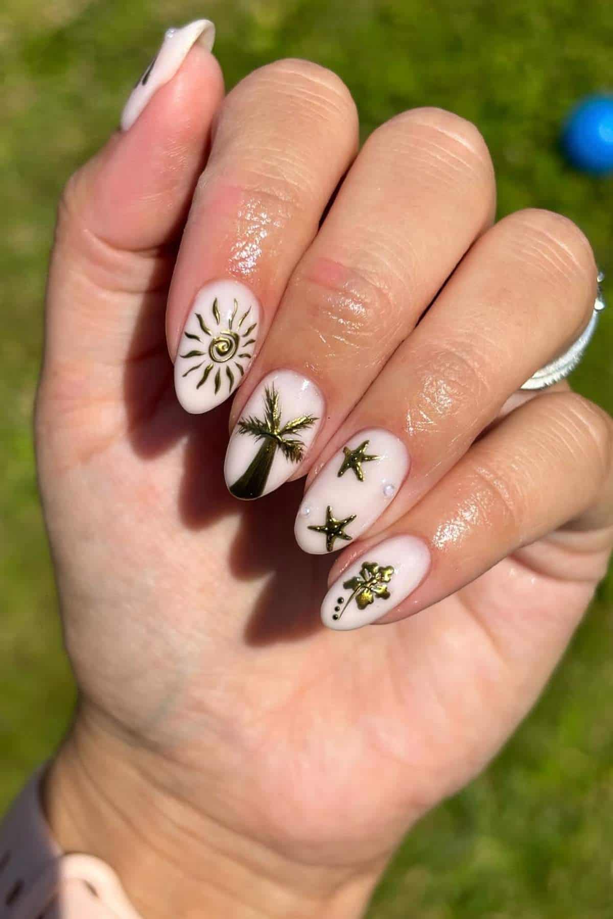 A hand with long nails painted light pink, featuring gold designs of a sun, palm tree, stars, and dotted accents. Grass and a blue object are visible in the blurred background.