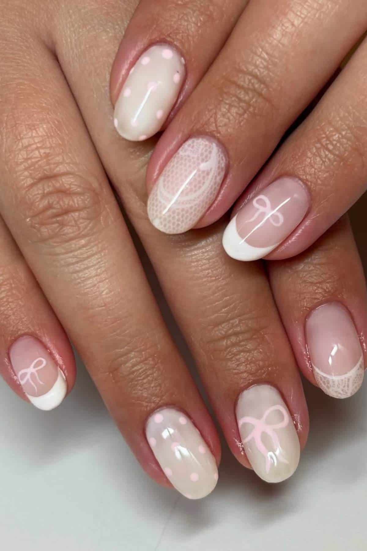 A hand with short, oval-shaped nails painted in a soft pink and white design, featuring bows, polka dots, and French tips.
