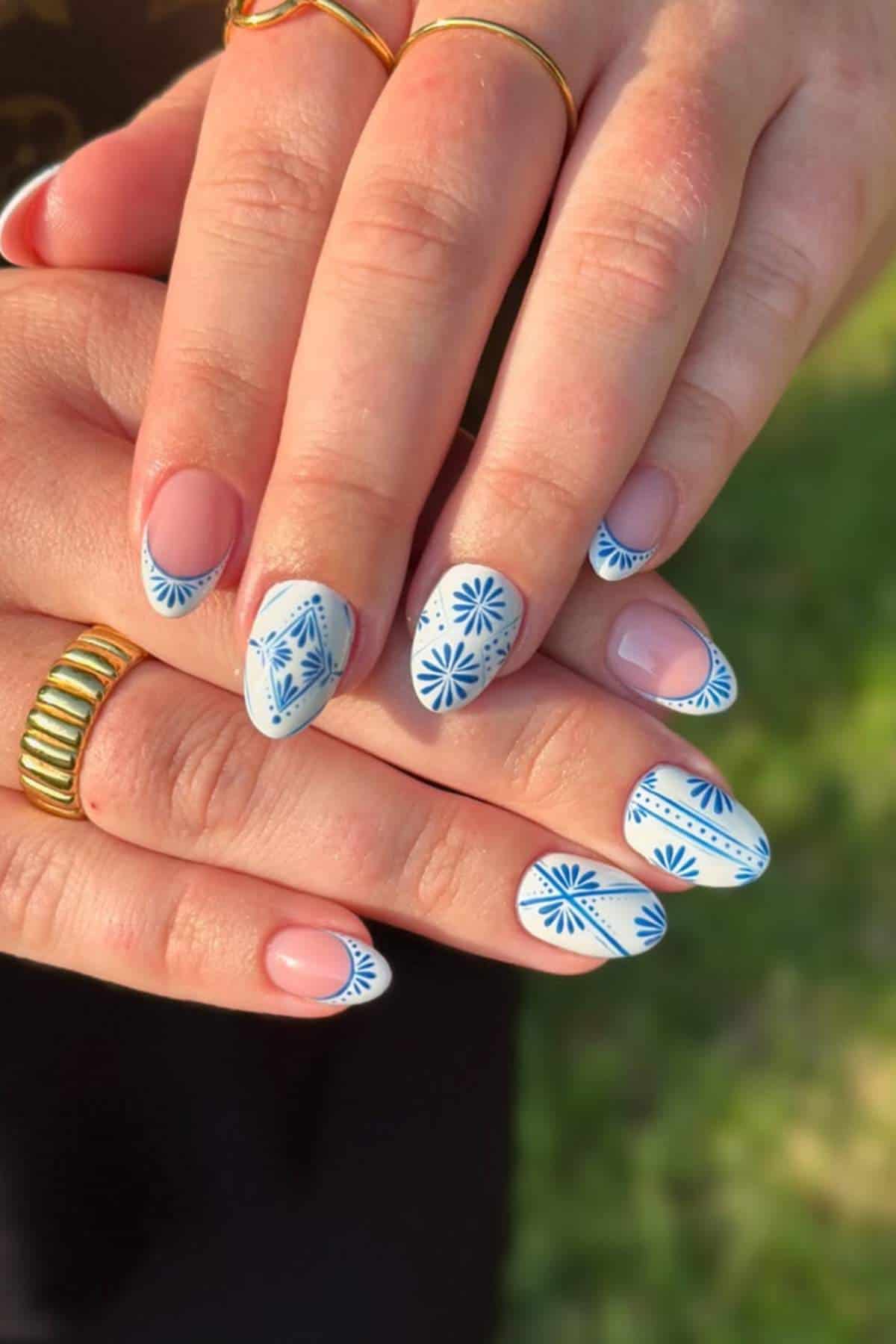 Hands with almond-shaped nails featuring blue floral and geometric designs on a nude base. The person is wearing gold rings.
