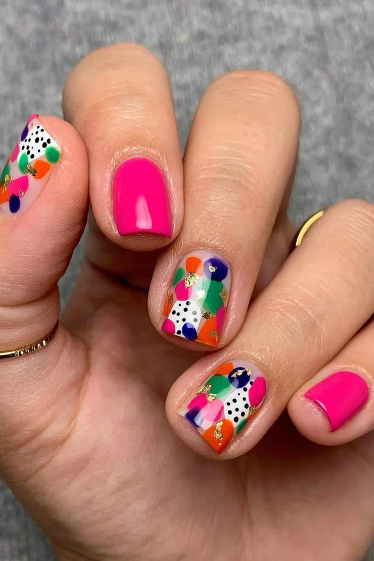 A hand with short nails; three nails are painted solid pink, and two nails have colorful abstract patterns with black and white dots.