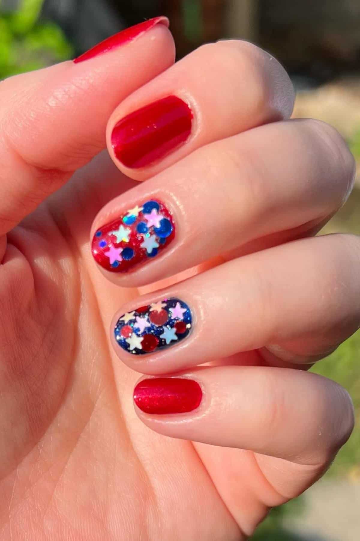 A hand with red nail polish on three fingers and multi-colored star-shaped glitter on the middle and ring fingernails.