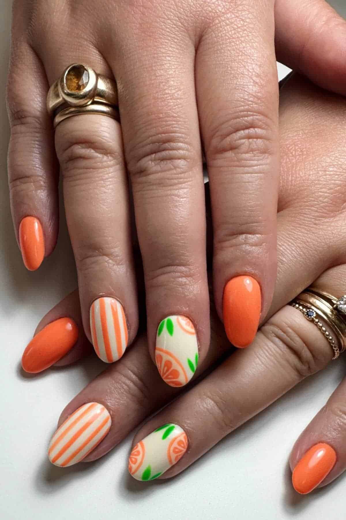 Hands with orange-themed nail art featuring solid orange nails, orange and white stripes, and citrus fruit designs. The person is wearing gold rings on their fingers.