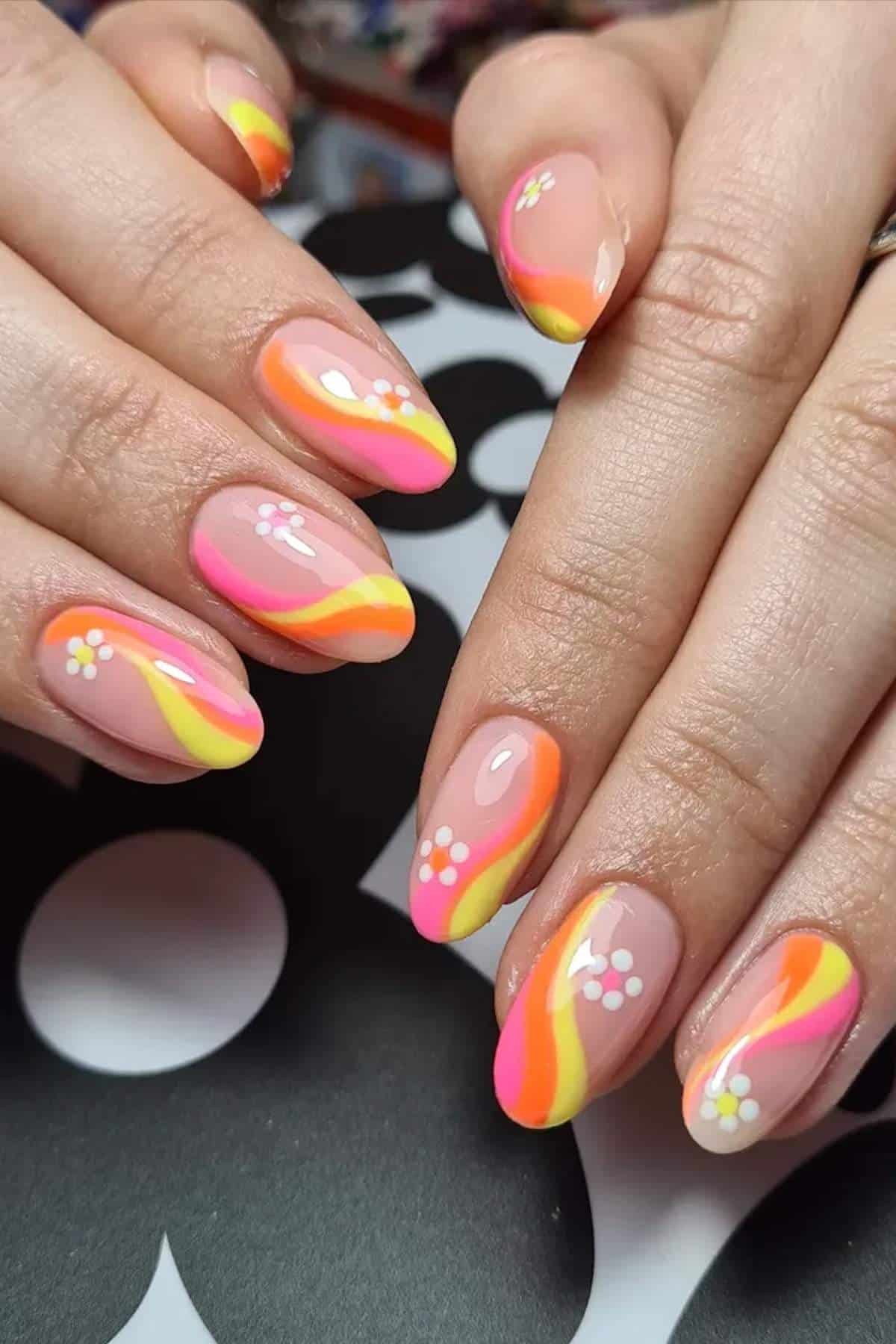 Hands with almond-shaped nails featuring pink, orange, and yellow wavy lines and small white daisy designs on a neutral base.