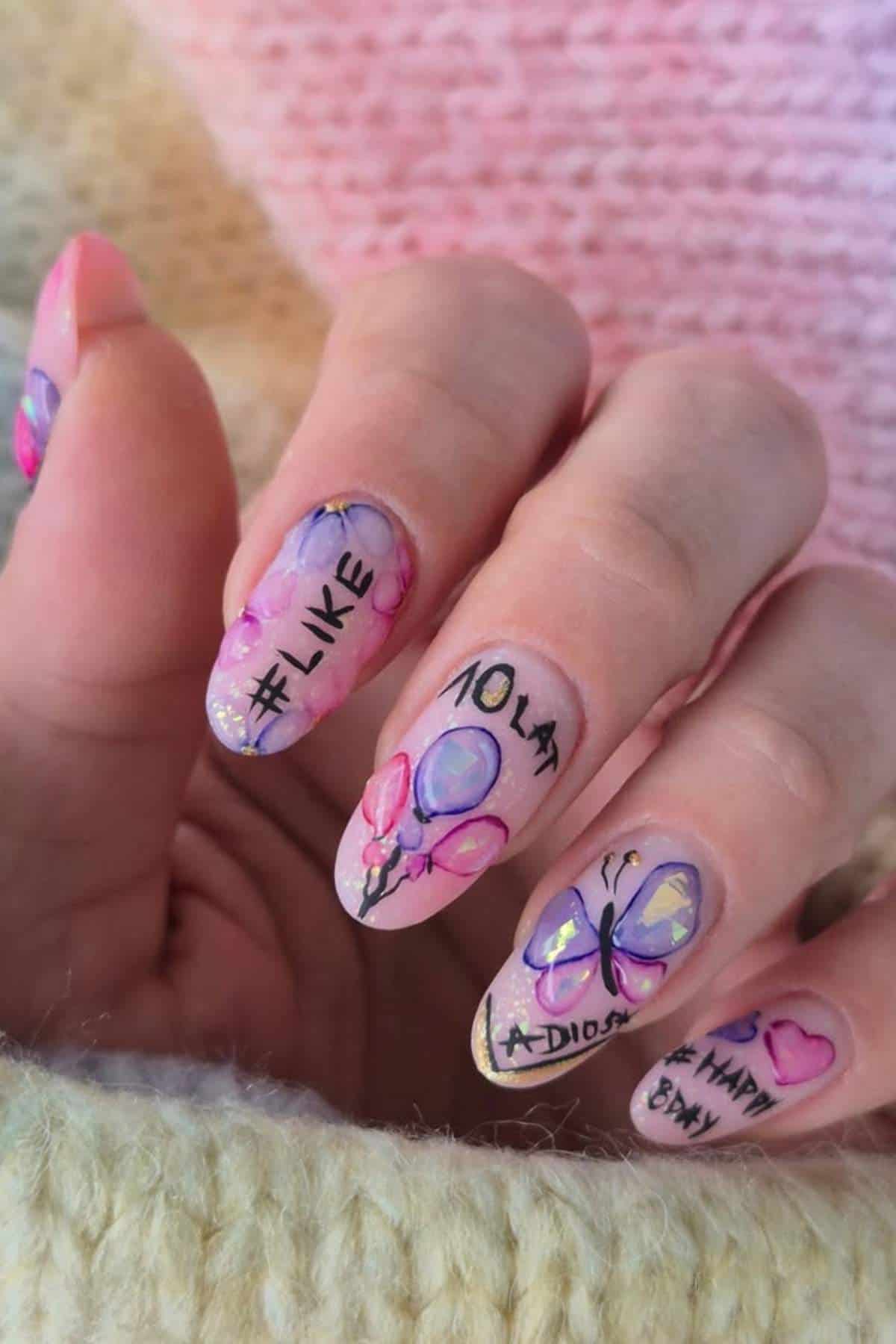 A hand with long, almond-shaped nails featuring colorful nail art with balloons, a butterfly, hearts, and words like "#LIKE," "LOLAT," "ADIOS," and "HAPPY BDAY.