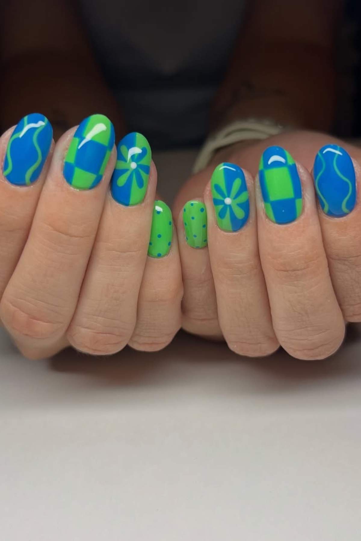 Hands with manicured nails featuring blue and green designs, including checkerboards, flowers, dots, and wavy lines.