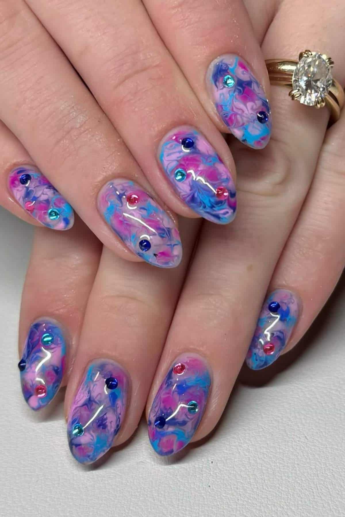 Hands with almond-shaped nails featuring a colorful marbled pastel design and small multicolored rhinestones. A gold ring with a large diamond is worn on one finger.