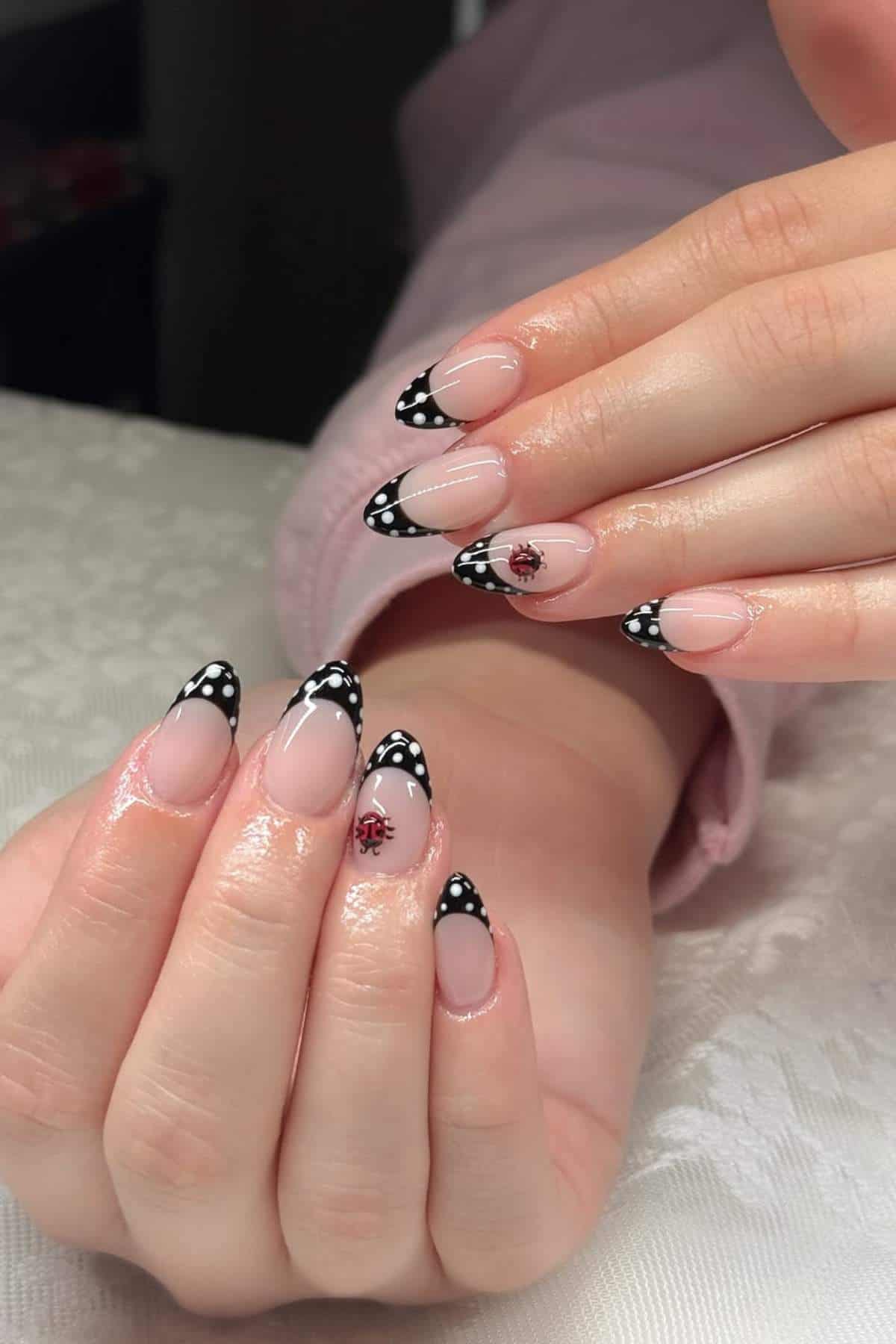 Close-up of hands with almond-shaped nails featuring black and white polka dot French tips and small ladybug designs on two accent nails.