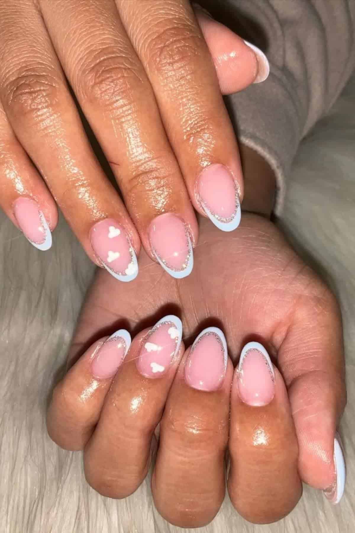 Hands with almond-shaped nails featuring a pale pink base, blue and silver outline tips, and white heart accents, displayed against a soft, white background.
