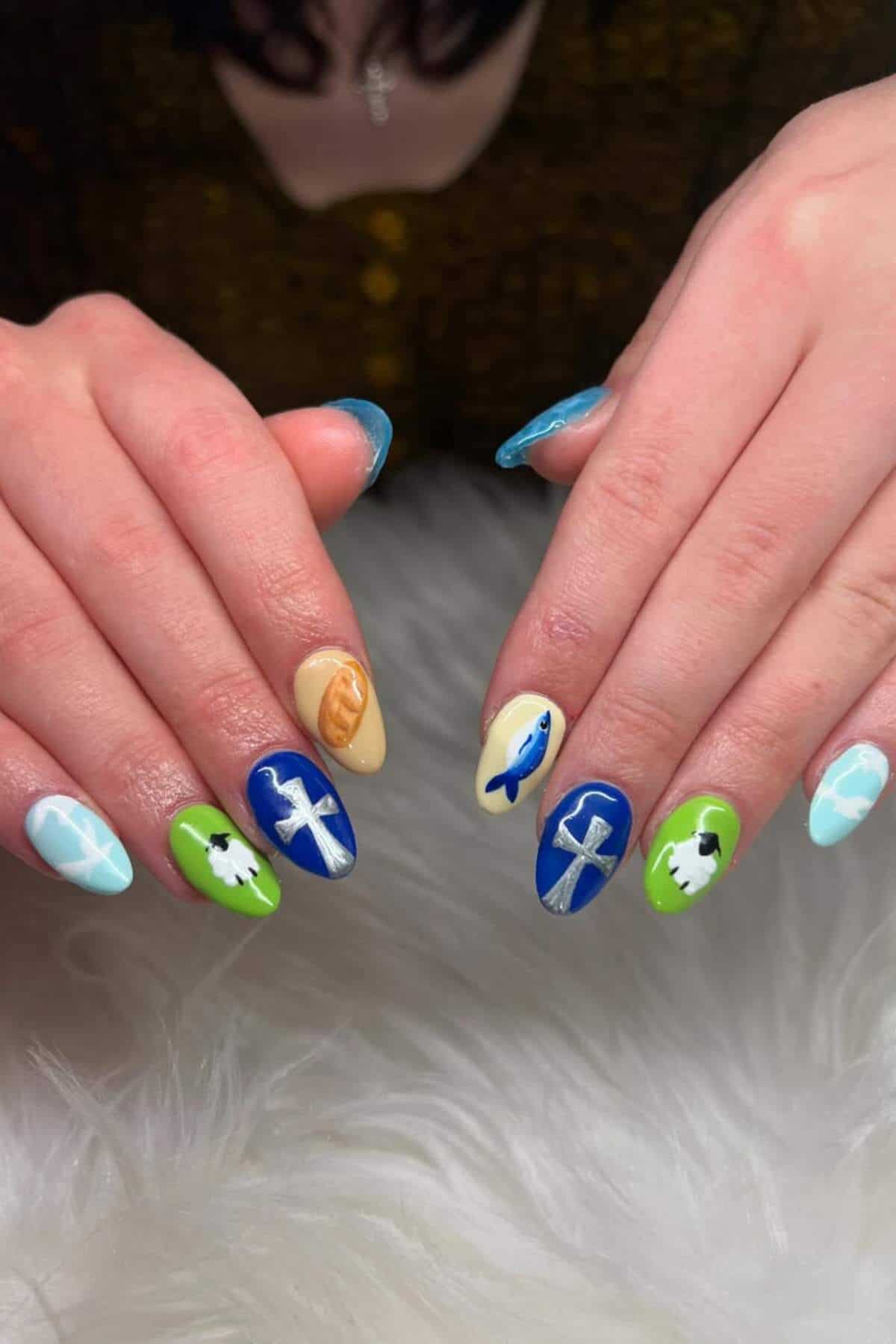 Hands with manicured nails featuring blue, green, and beige polish with designs of clouds, sheep, fish, crosses, and bread on a white textured surface.