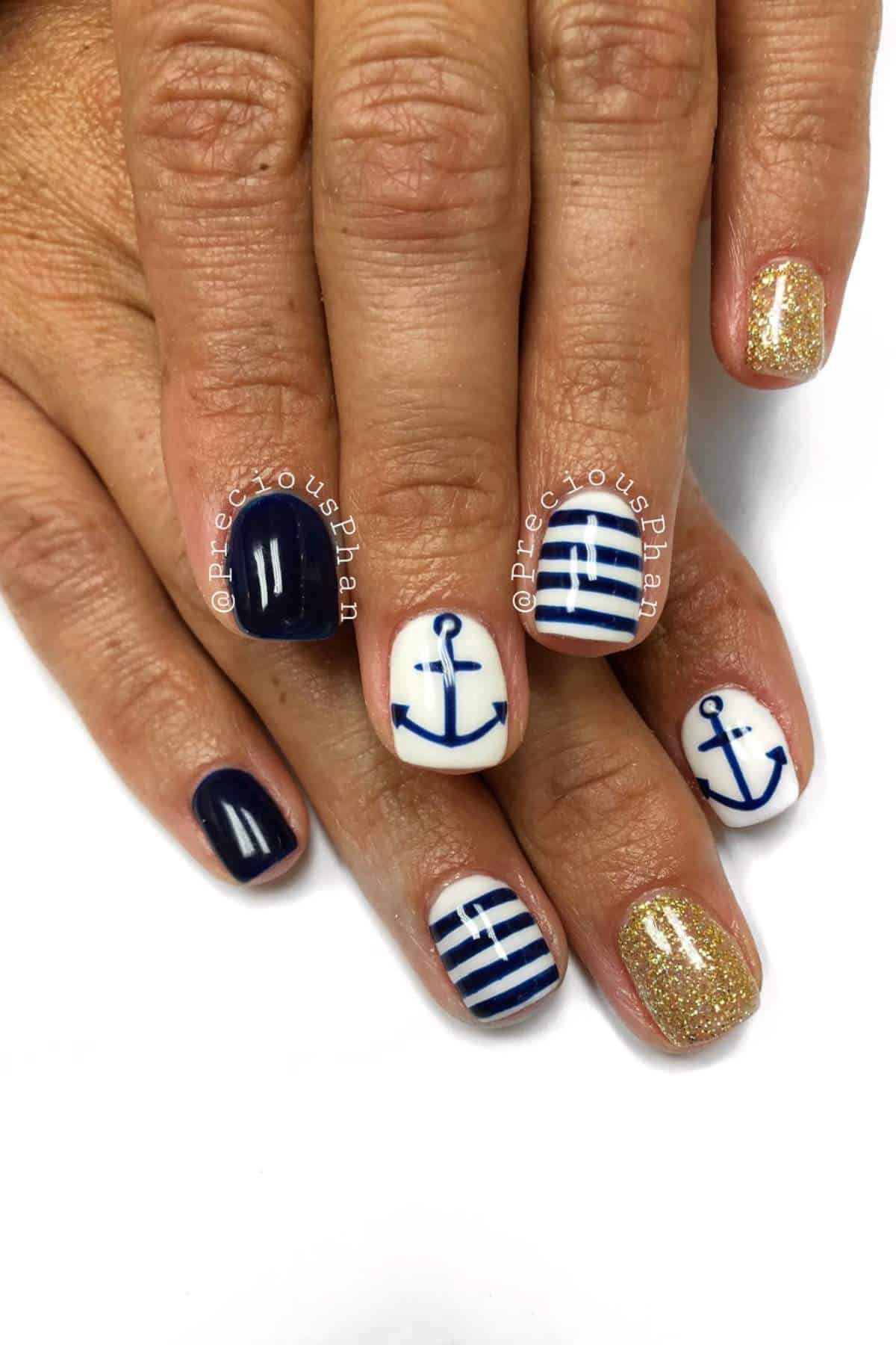 A hand with nautical-themed nail art featuring navy blue polish, gold glitter, white anchors, and blue horizontal stripes on alternating fingernails.