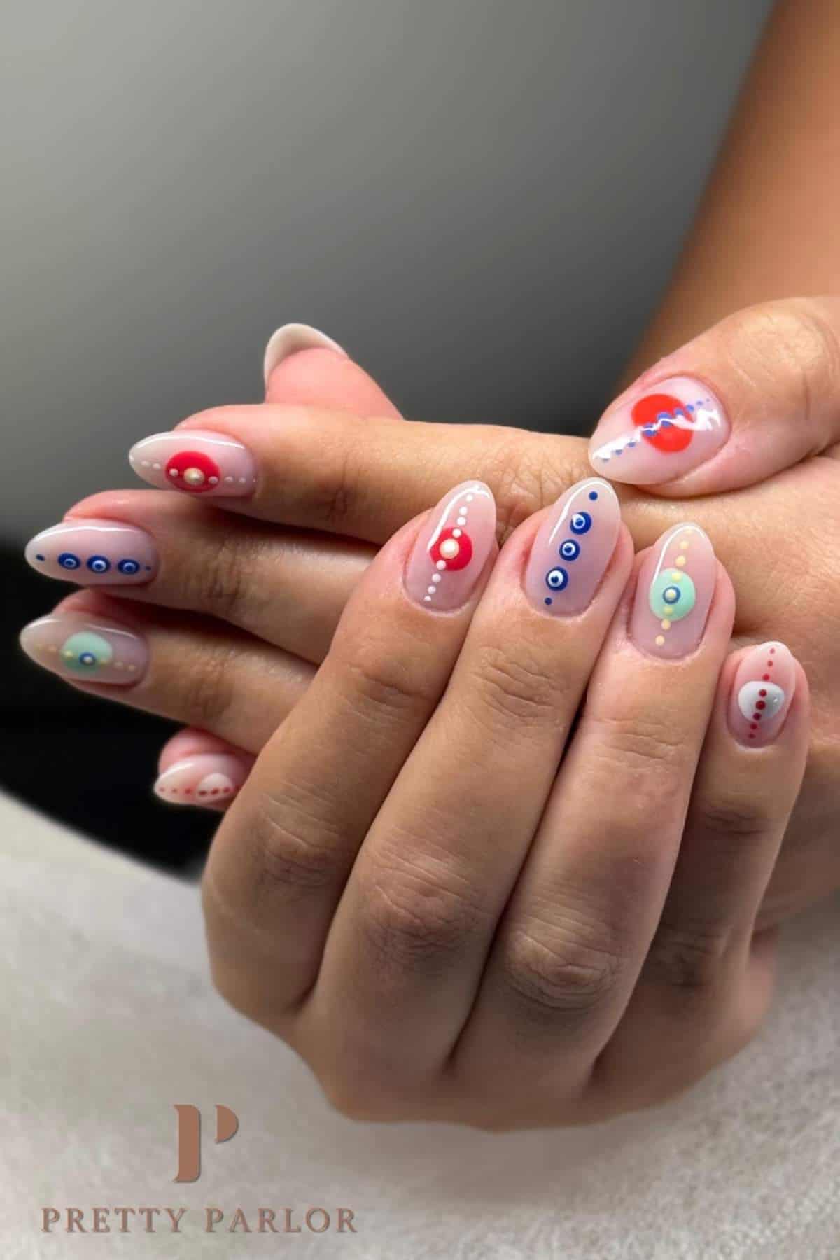 Hands with almond-shaped nails featuring a nude base and colorful abstract dot designs in blue, red, and green. Logo in the corner reads "Pretty Parlor.