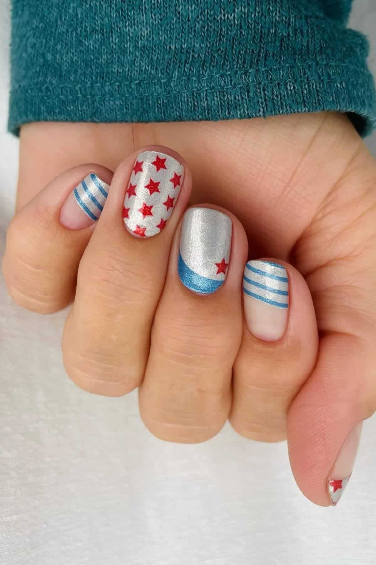 A hand with short nails painted with a patriotic design featuring red stars, blue stripes, and metallic silver and blue accents.