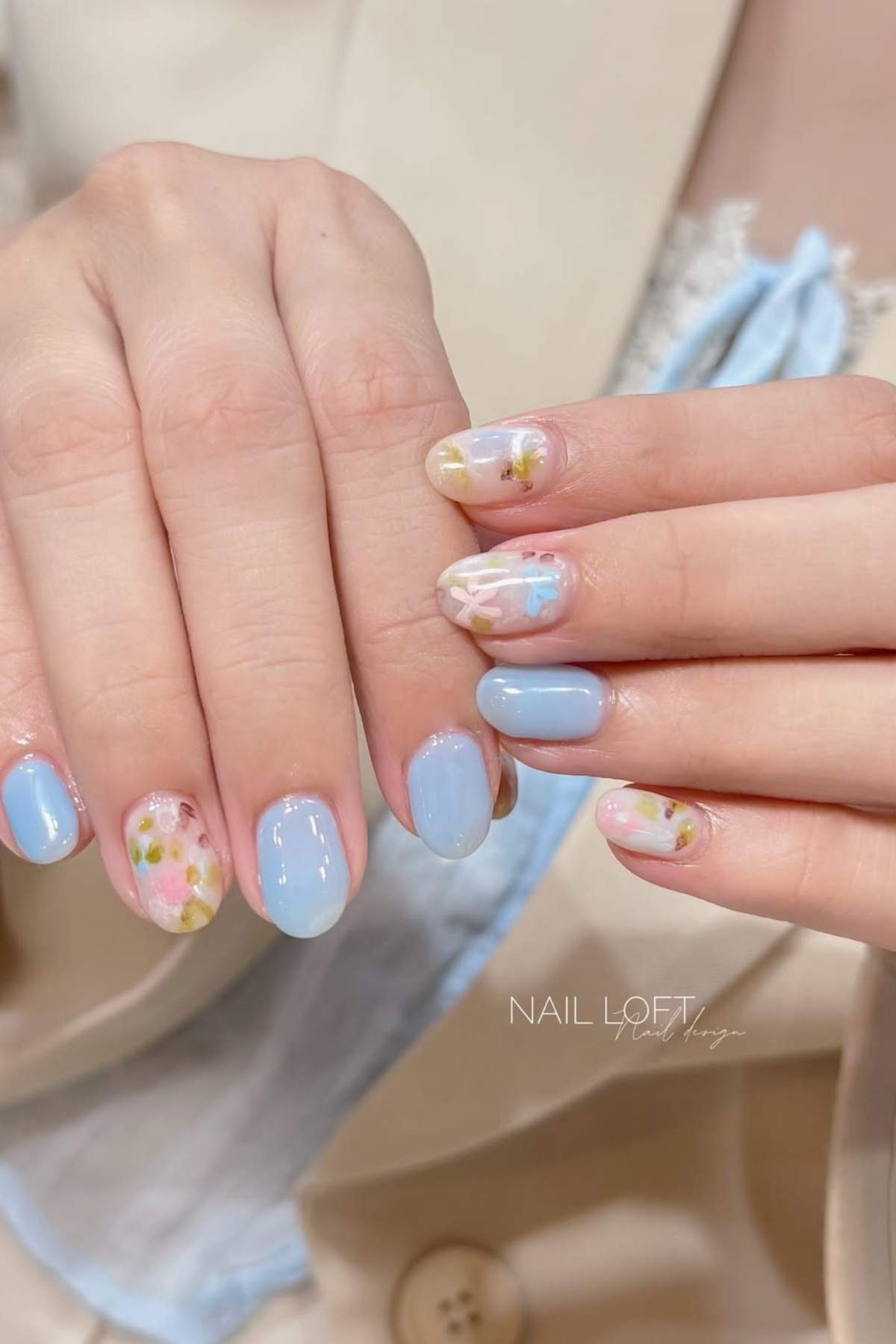 Hands with pastel blue nail polish and accent nails featuring floral and geometric designs. The person is wearing a beige jacket with a light blue top underneath.