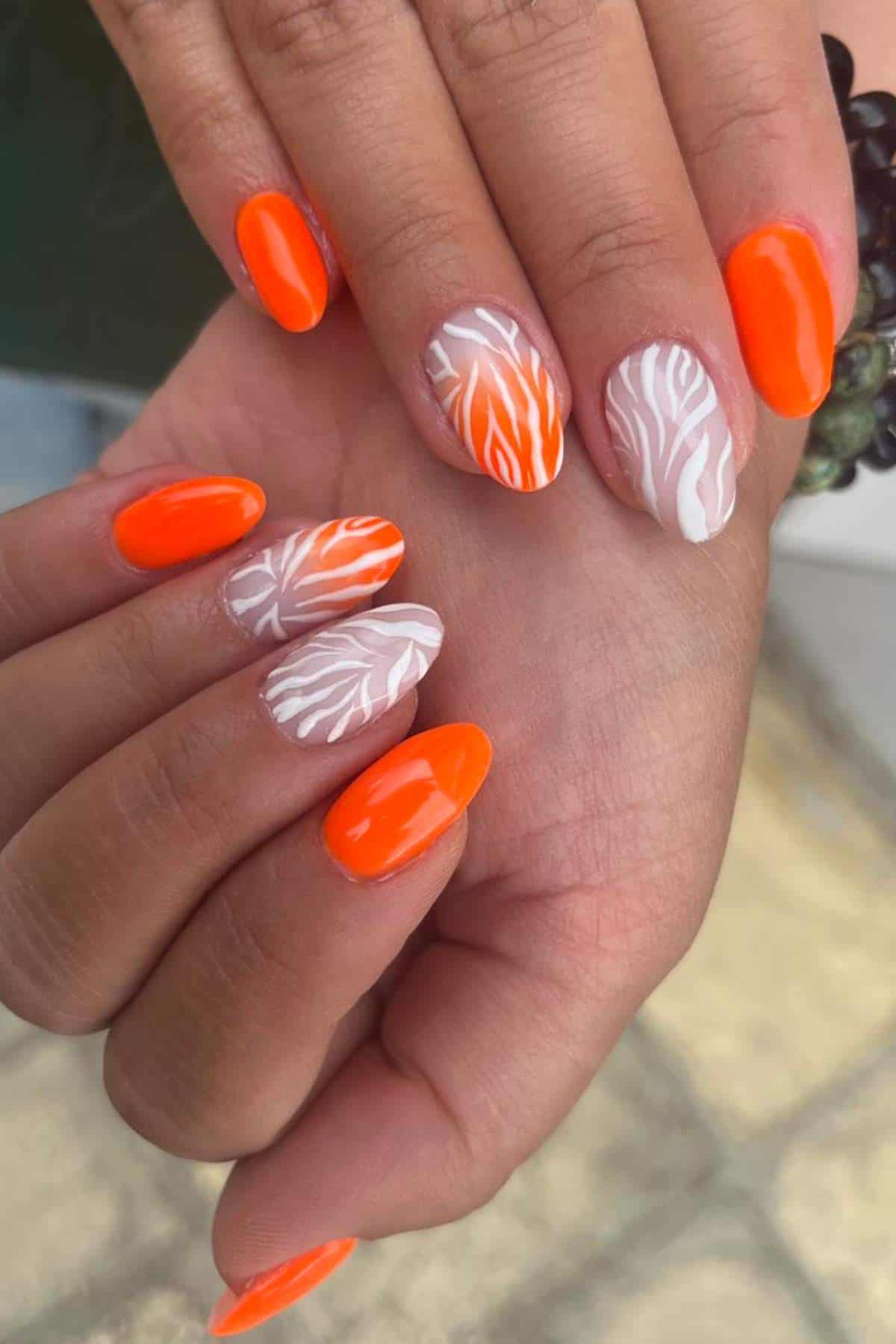 Hands with oval-shaped nails painted in bright orange and nude polish, featuring white leaf designs on some nails.